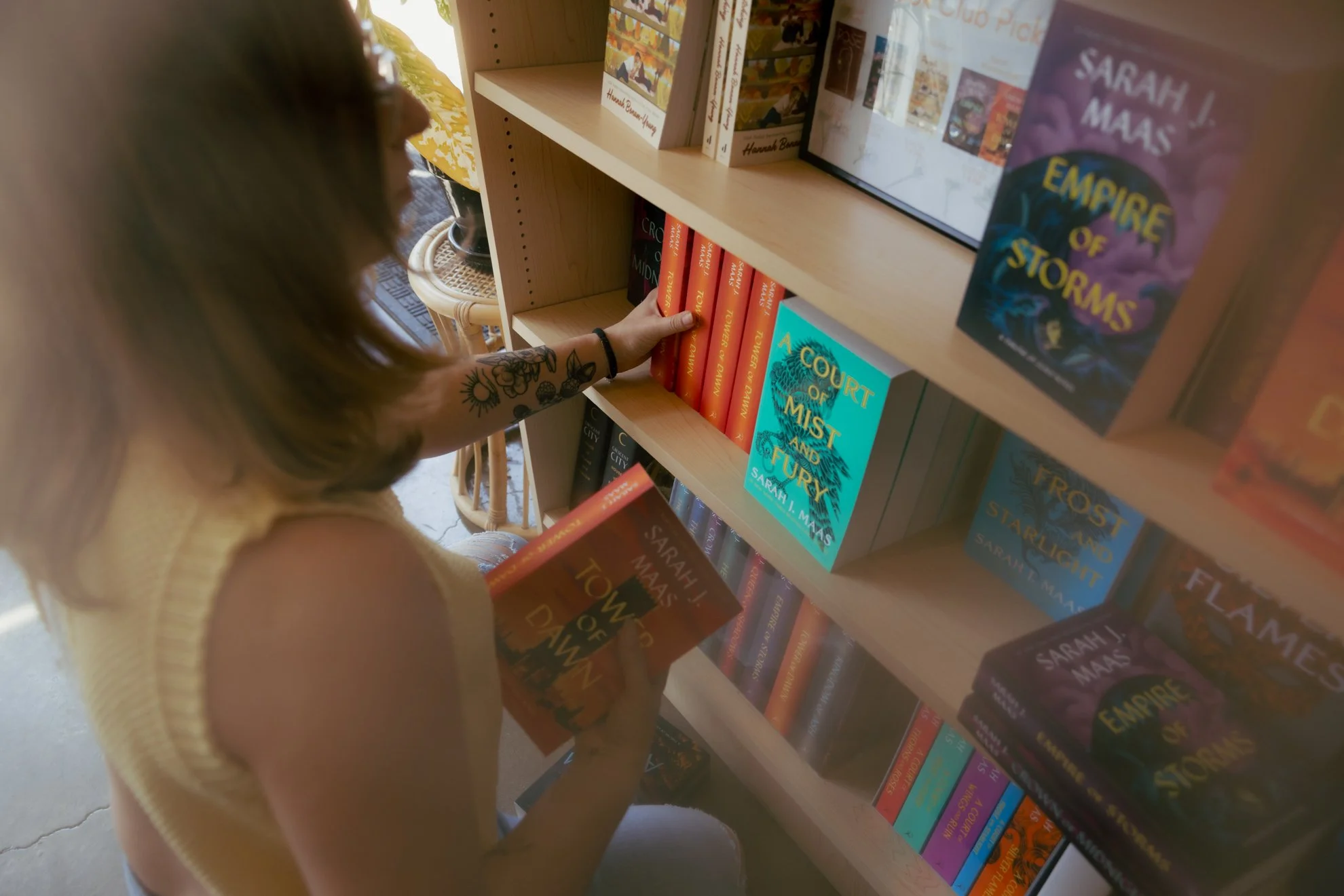 A person in a yellow sweater with tattoos on her arm is browsing books on a bookshelf in a bookstore. She is holding a book titled 'Towers of Rain' by Sarah J. Maas, with other books visible on the shelf.