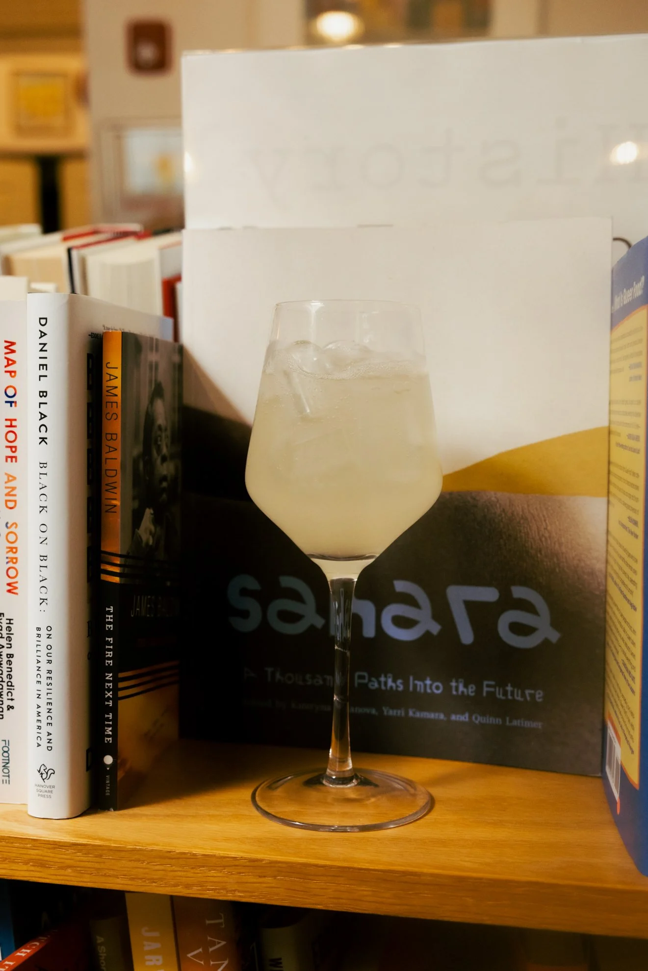 A glass of white wine with ice on a wooden shelf surrounded by books.