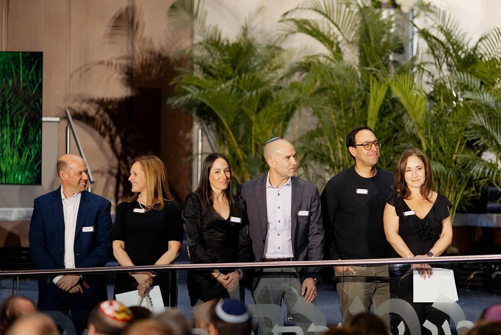 Group of six people standing on a stage, dressed in business attire, with large green plants and a wood-paneled wall in the background. They are facing slightly to the right, engaged in an event or presentation.