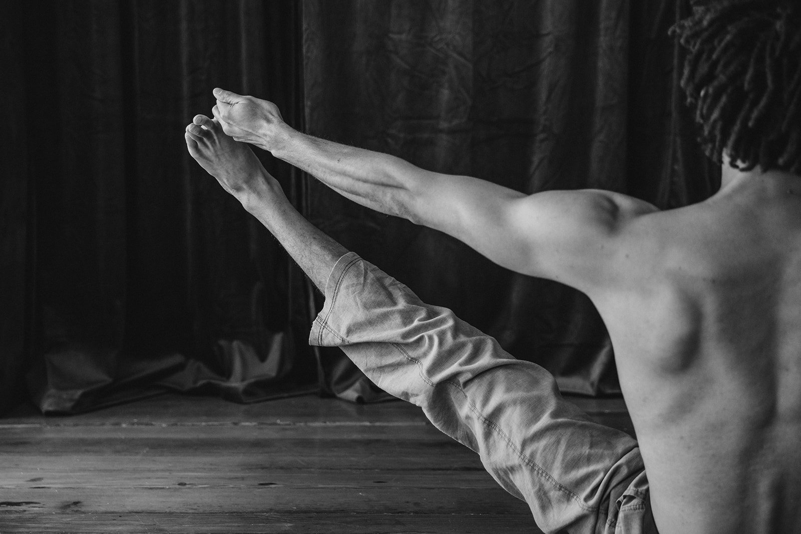 A person doing yoga or stretching on the floor, with one arm extended forward and the other stretched back, shirtless and wearing jeans, against a dark background.