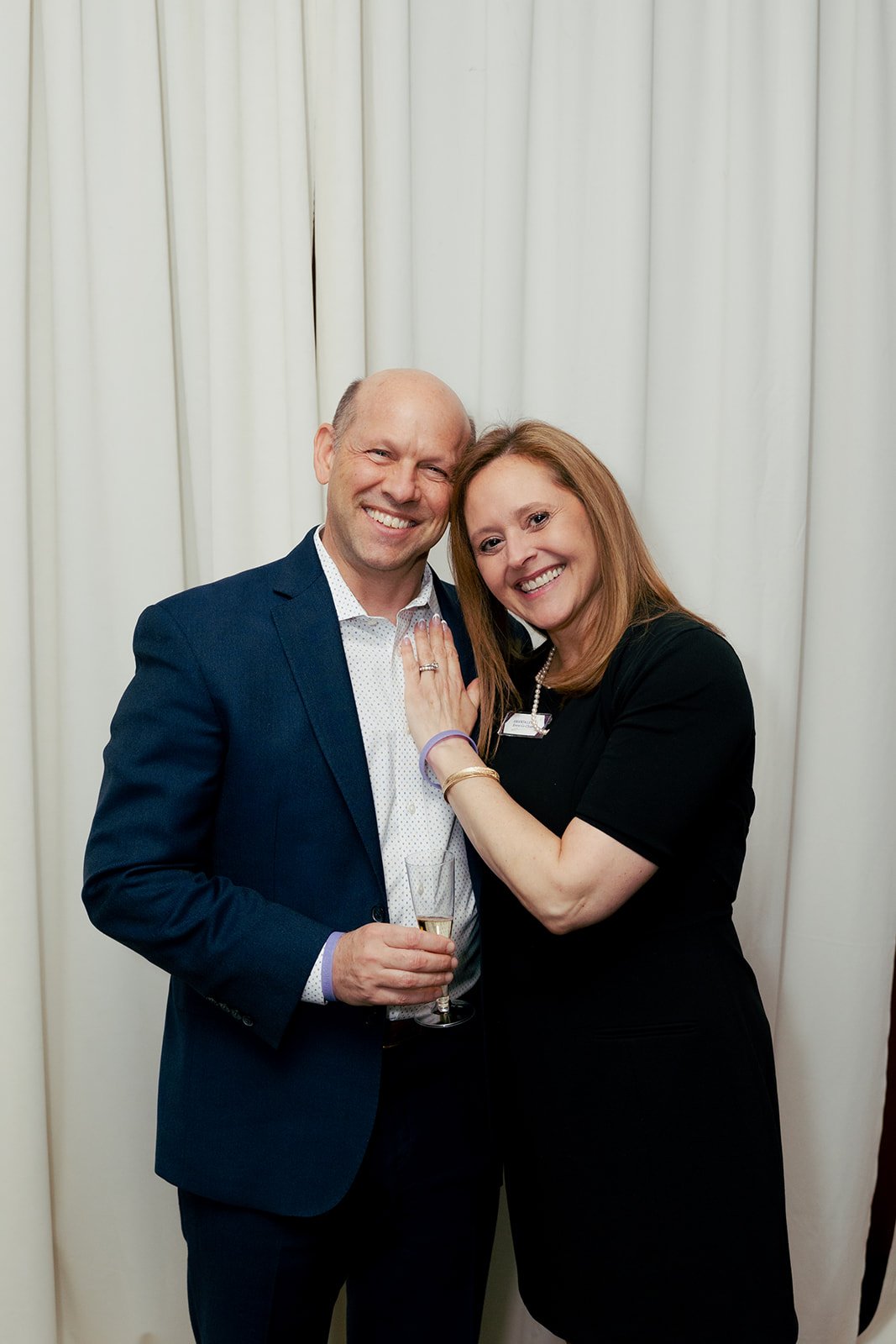 A man and a woman smiling and posing together in front of a white curtain. The man is wearing a dark suit and holding a glass of champagne. The woman has red hair, wears a black dress, and a name tag. She has her hand on the man's chest, showing a ring on her finger.