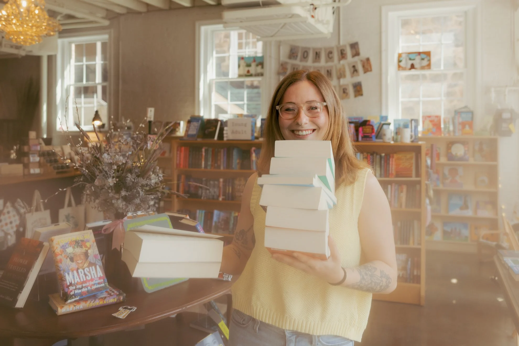 A smiling woman with glasses and tattoos on her arm, standing behind a table in a bookstore, holding a stack of books. The background shows bookshelves, a banner reading 'HOME', and a flower arrangement.