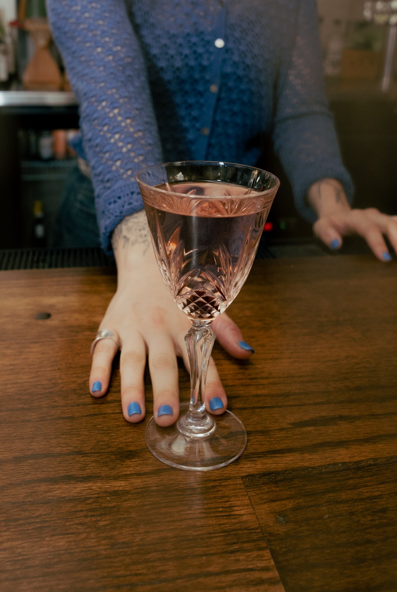 A person with blue painted nails reaching out with a hand on a wooden table, holding a glass of pink wine, with another person in a blue knit sweater leaning over the table.