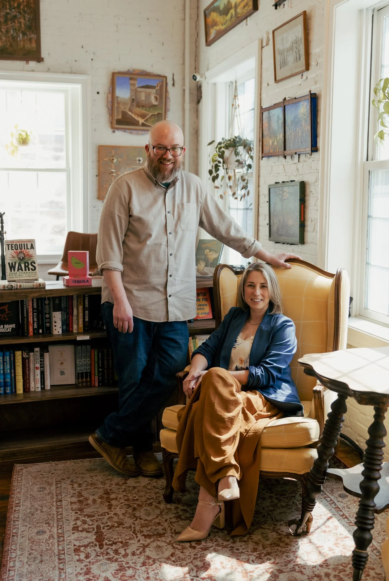 A man with glasses and a beard stands next to a seated woman with blonde hair in a room with white brick walls, bookshelves, and framed paintings, near large windows with sunlight.