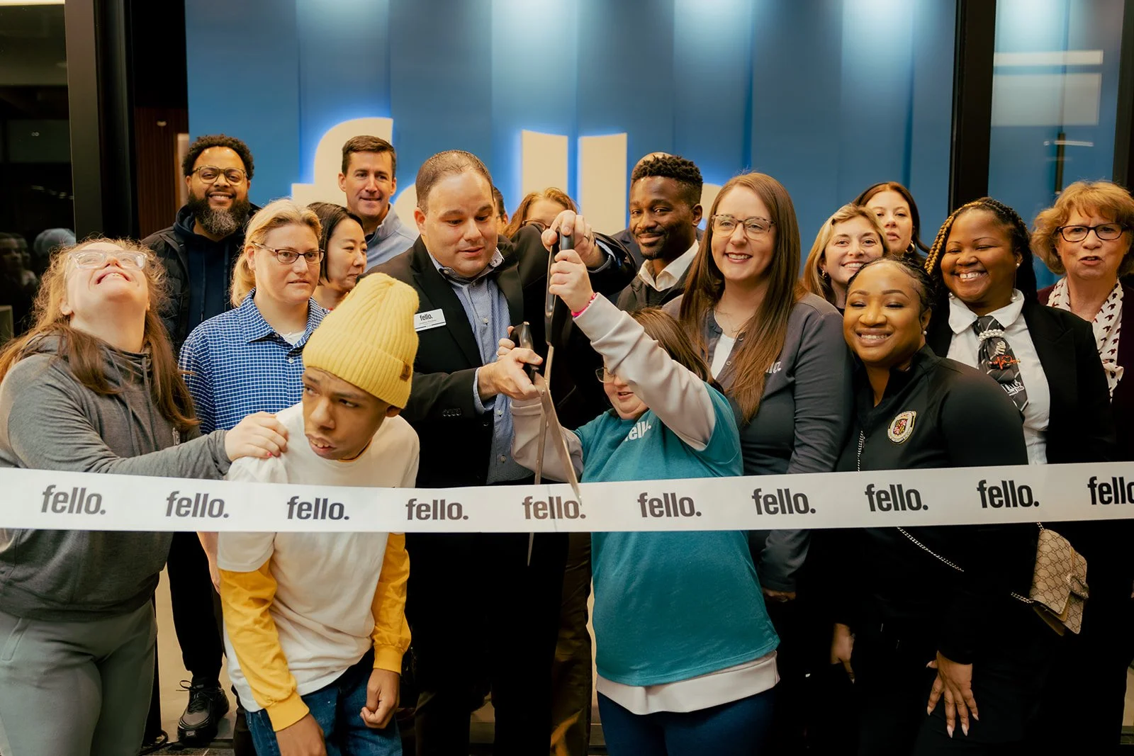 A diverse group of people gathered at a ribbon-cutting event, with some holding scissors, in front of a blue background with the 'Fello' logo repeated on the ribbon.