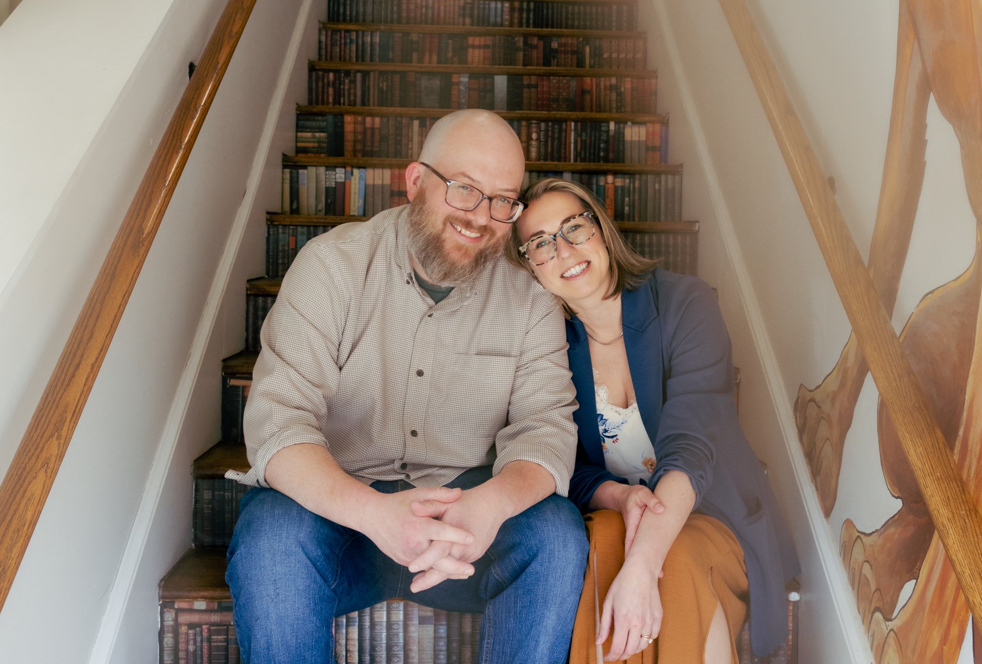 A smiling man and woman sit on a staircase with a bookshelf behind them. They are leaning close to each other, wearing glasses, and looking at the camera.