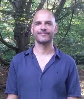 Man with a shaved head and goatee smiling, standing outdoors with trees in the background.