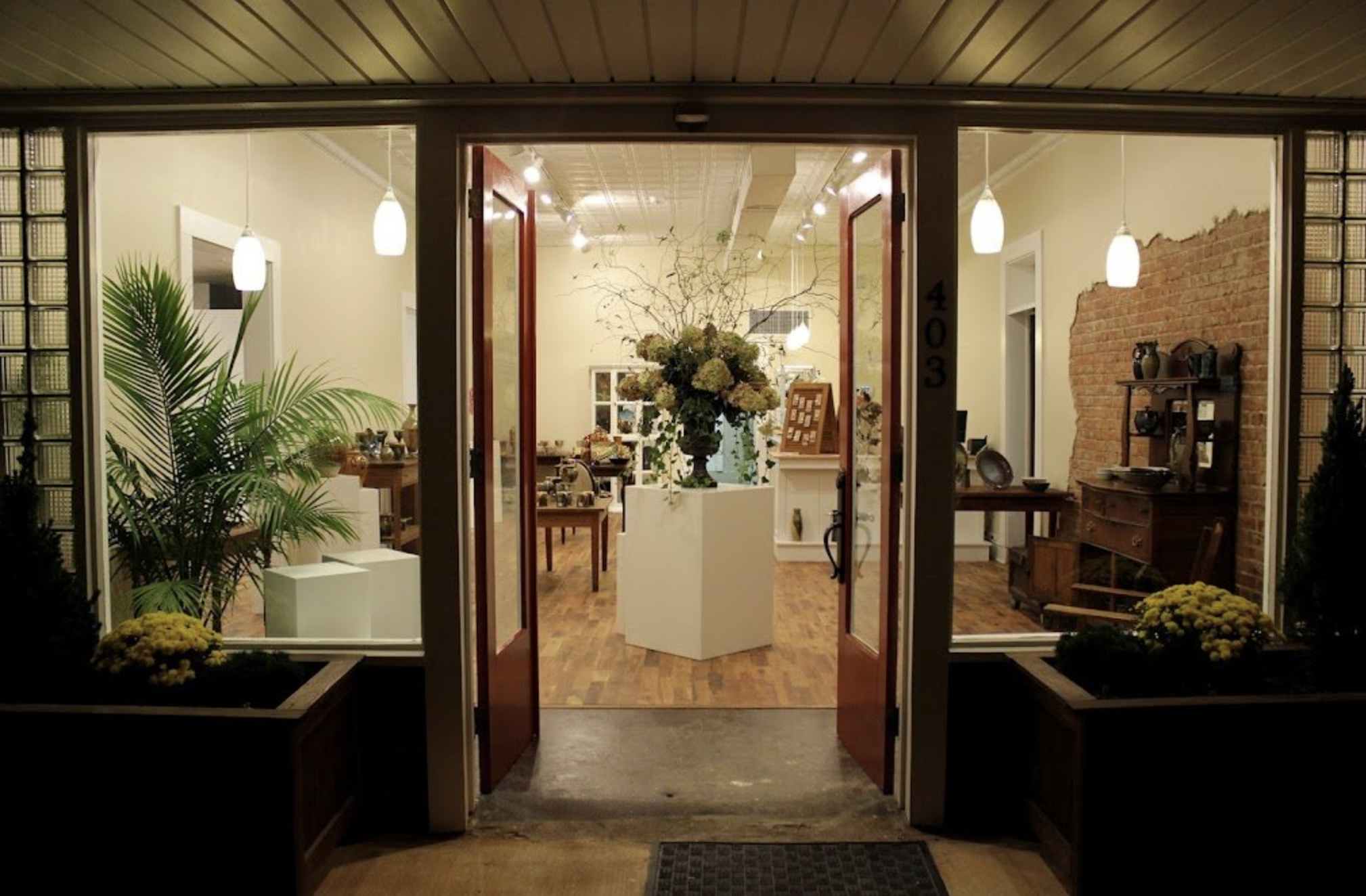 Open front door looking into a warmly lit gallery in Morganton, NC area with plants, a large floral arrangement, and rustic brick wall.