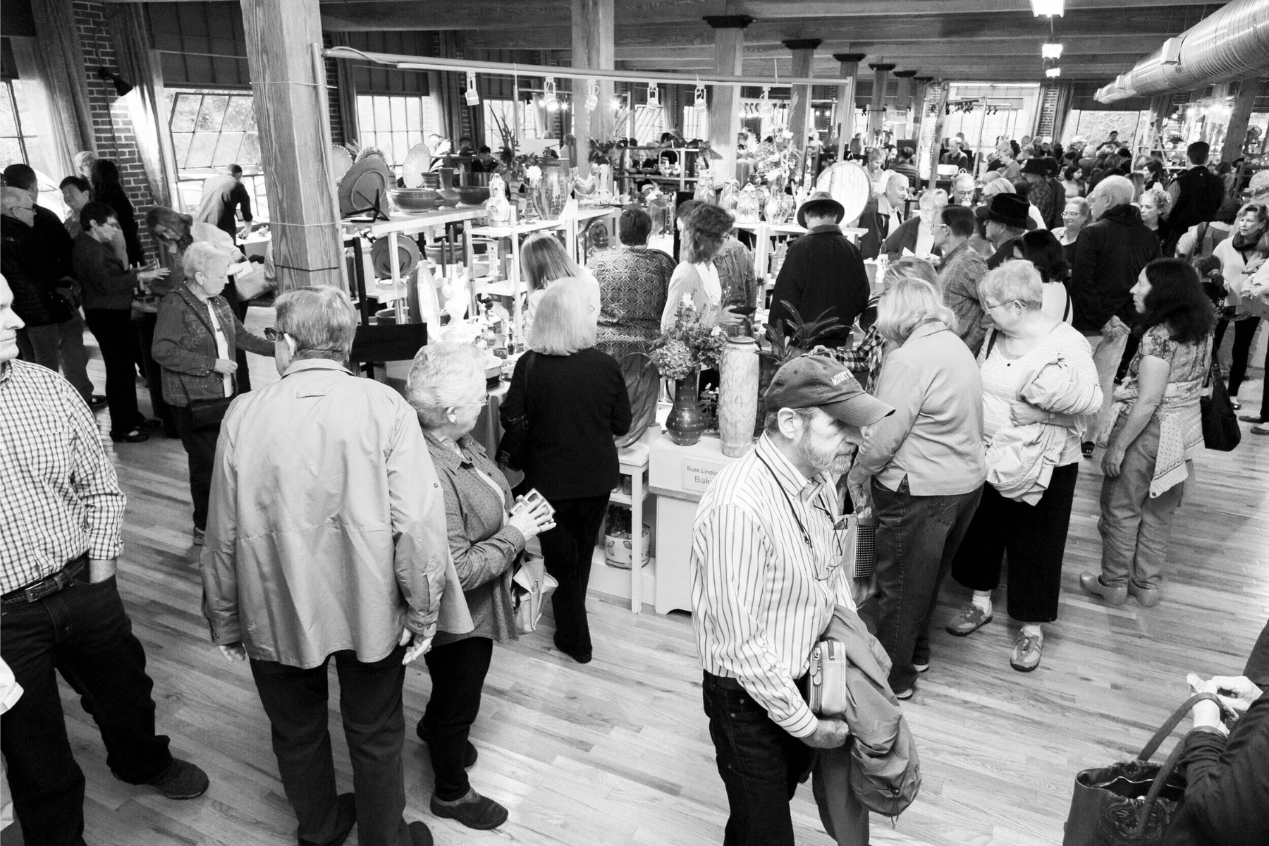 People shopping indoors at the annual Spruce Pine Potters Market, browsing and walking around various display tables and booths featuring pottery, vases, and decorative items.
