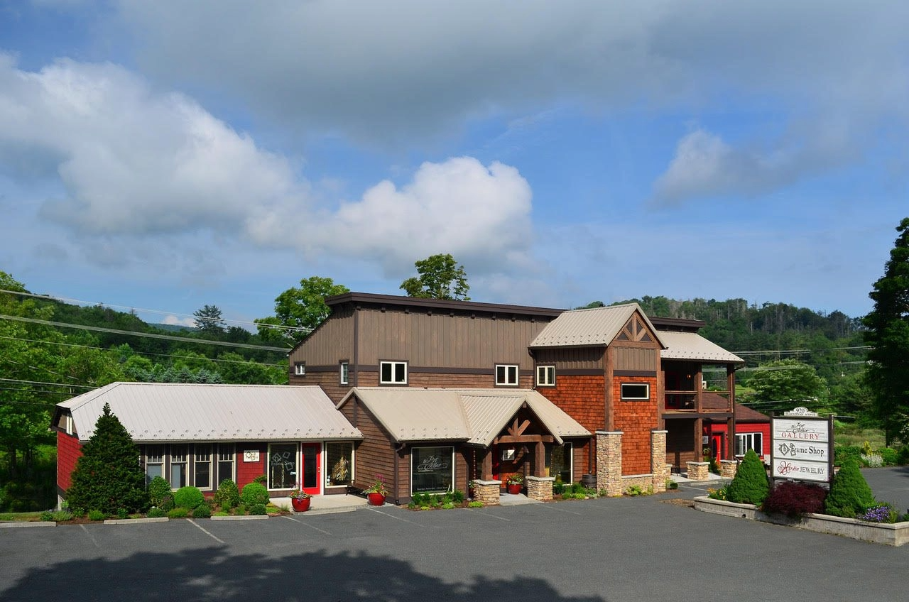 The multi-story building of the Art Cellar Gallery with a mix of wood and brick exterior, situated next to a parking lot, with greenery and trees in the background under a partly cloudy sky.