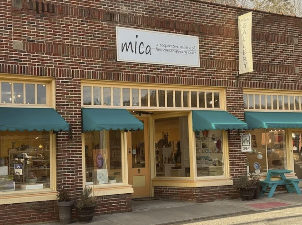 Front of a brick building with signs for 'mica' and 'GALLERY,' featuring large display windows with teal awnings, potted plants, and a turquoise outdoor bench in Bakersville, NC.