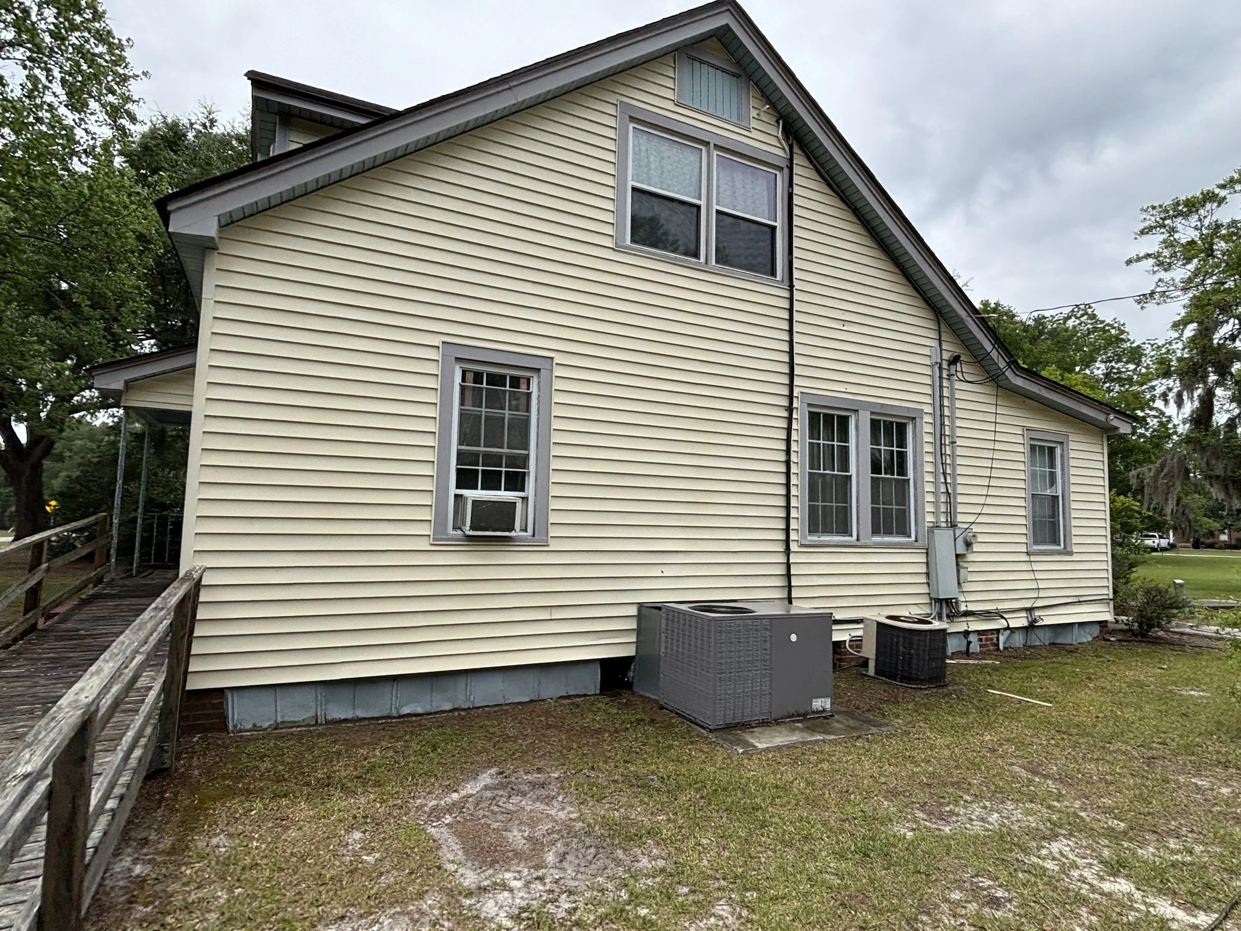 The image shows the side of a yellow house with white trim and multiple windows. There is a small wooden ramp leading up to the house and outdoor air conditioning units on the ground. The yard has some grass and dirt patches, and trees are visible in