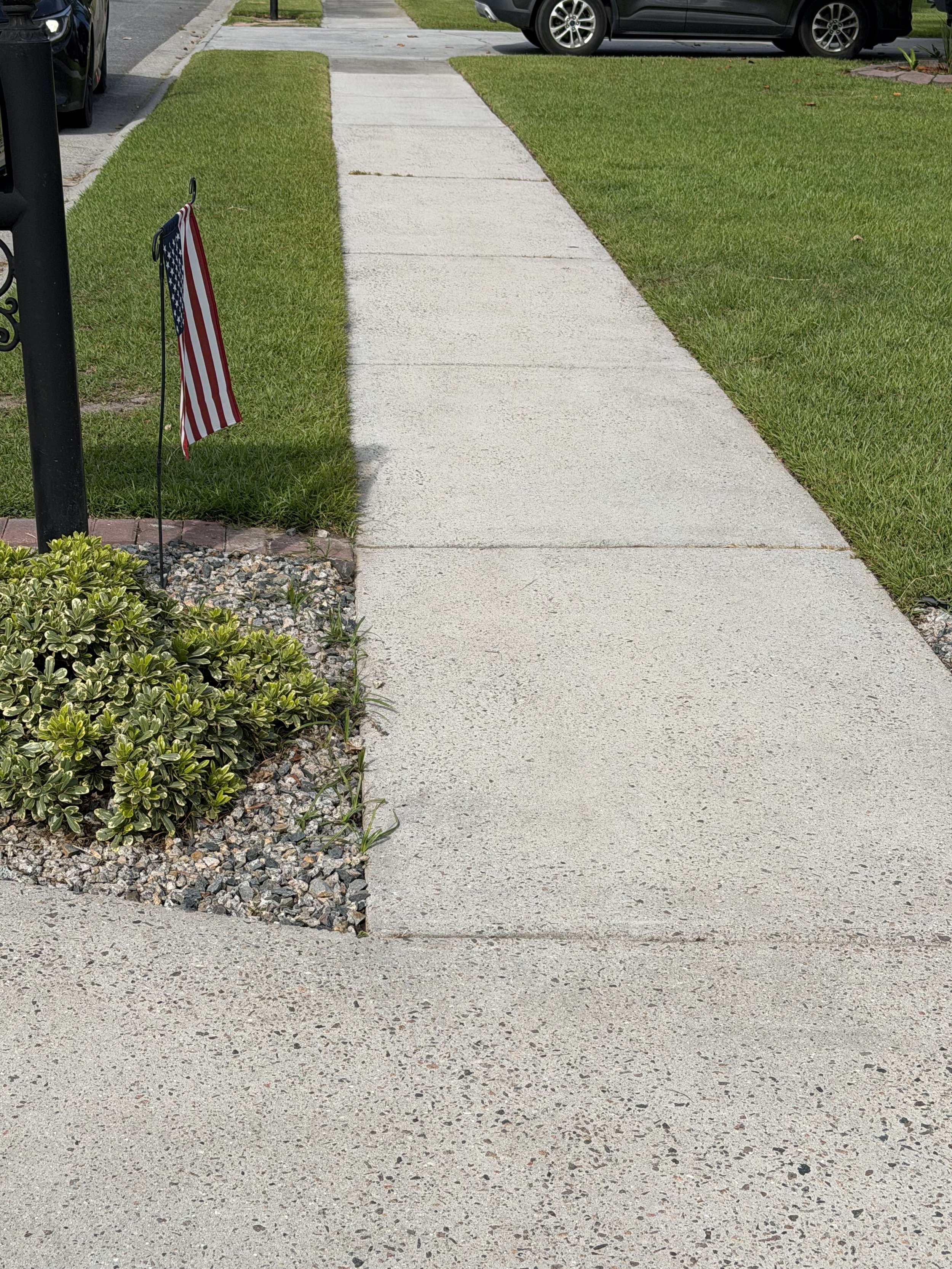 An American flag on a small flagpole next to a grassy area and a concrete sidewalk. In the background, there are cars parked along the street and part of a black mailbox is visible to the left.