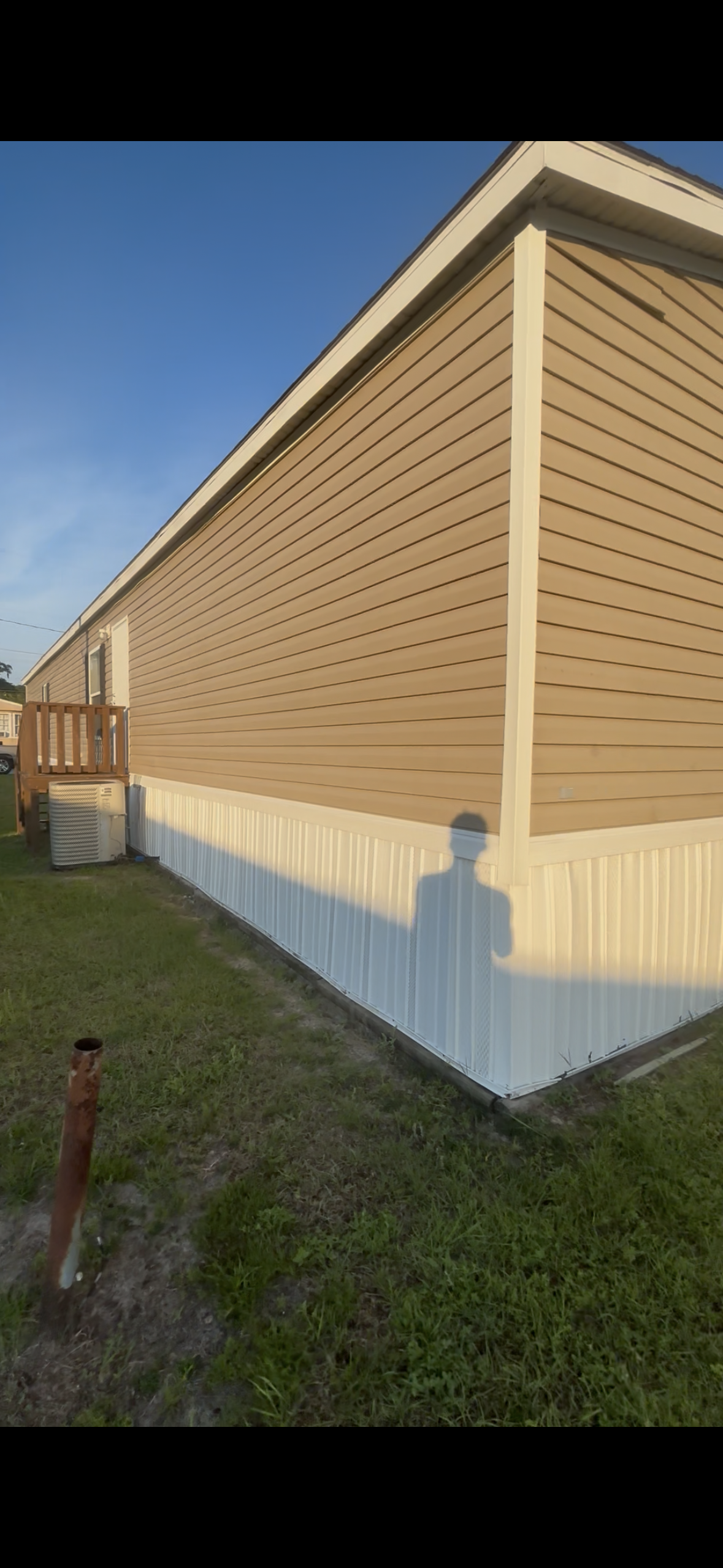 Side view of a mobile home with beige vinyl siding and white skirting, a small wooden deck with stairs, nearby HVAC unit, and the shadow of a person on the ground during sunset.