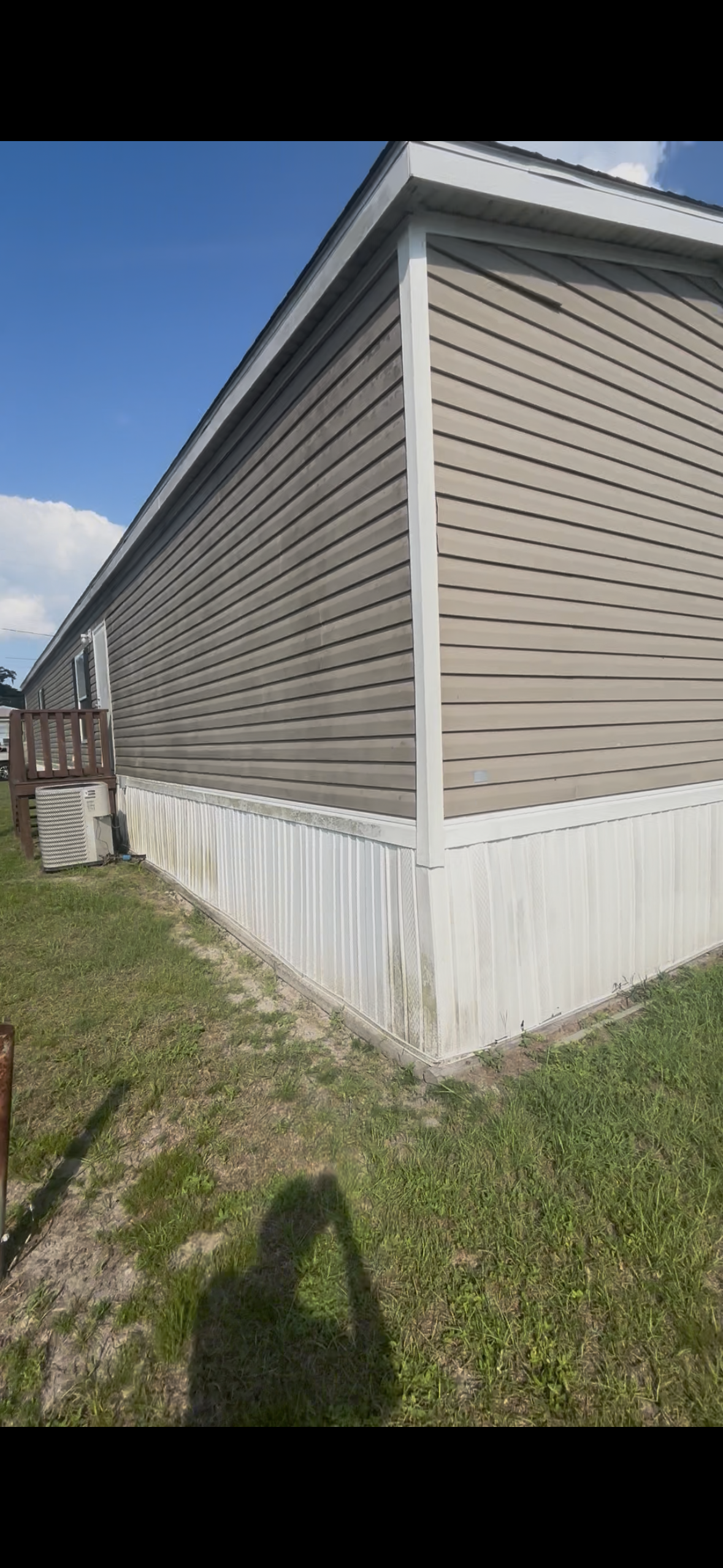 Side view of a beige mobile home with vinyl siding, a small wooden staircase, and an air conditioning unit outside. The ground is grassy, and the sky is blue with some clouds.