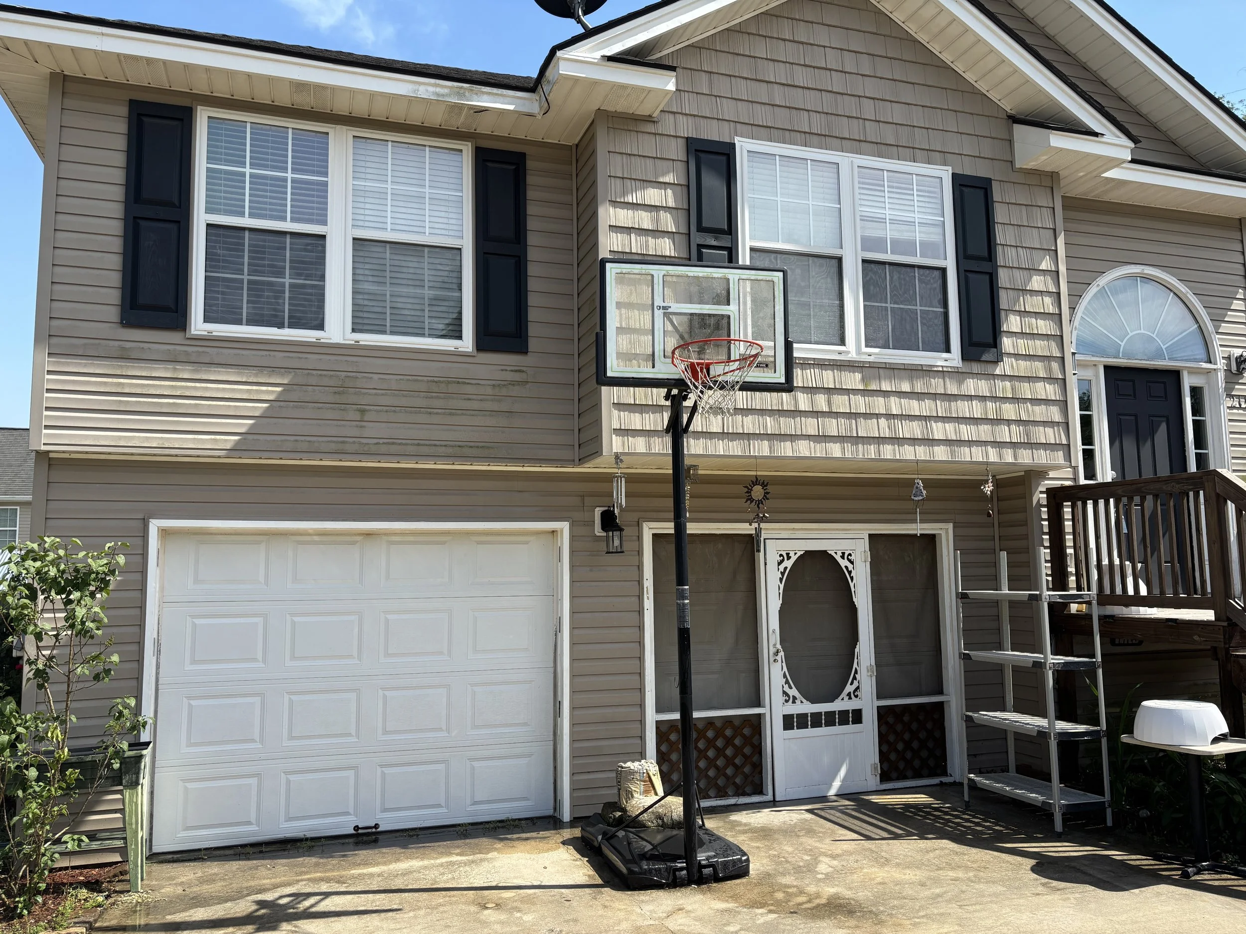The second story of a beige house with white trim and black shutters, featuring a basketball hoop placed in front of a window. The house has a two-car garage with a white door and a sliding glass door next to a decorative white gate. There are some p