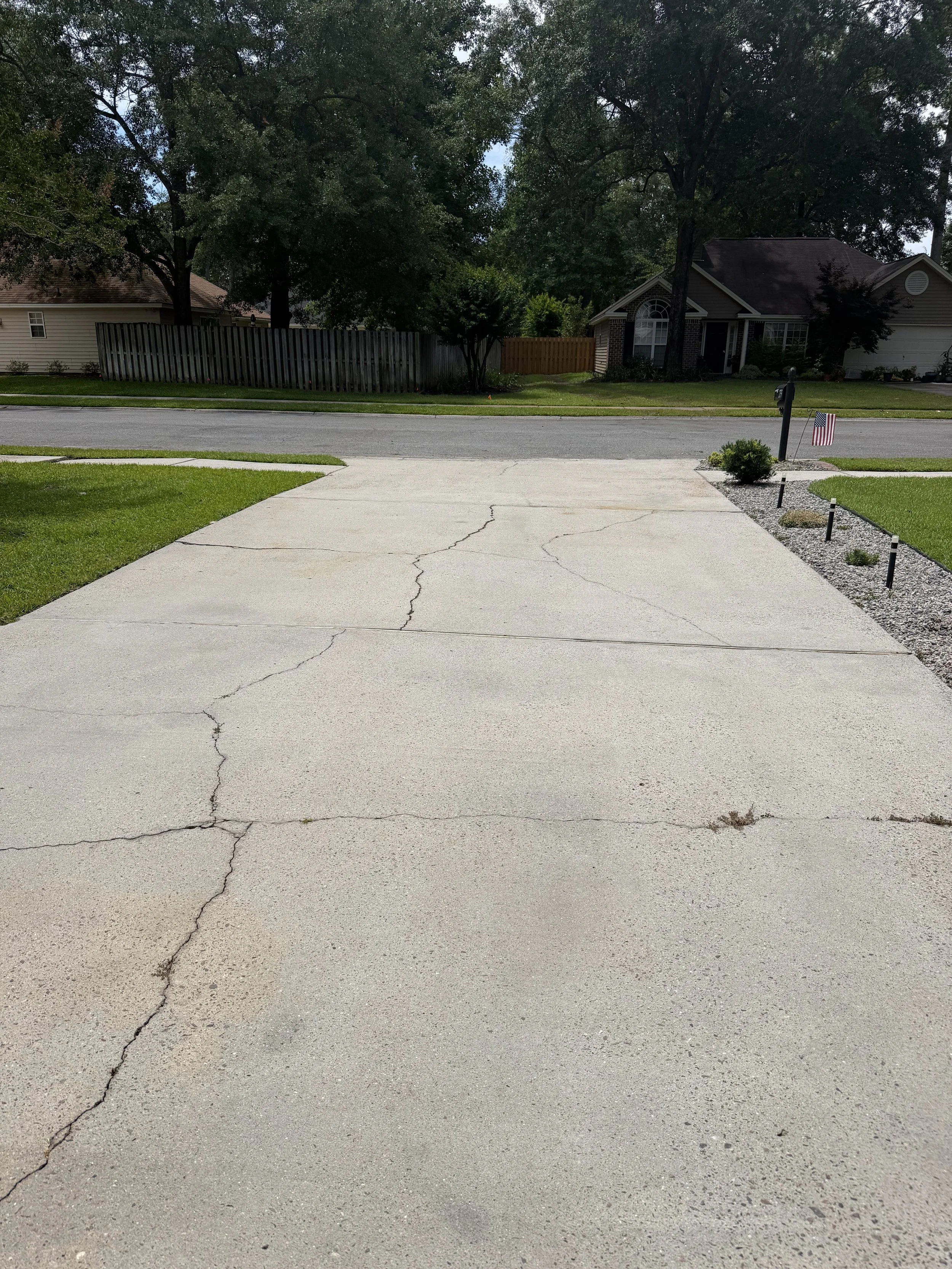 View of a suburban neighborhood with houses, trees, a lawn, and a concrete driveway with cracks. A mailbox with an American flag and small decorative posts are visible near the driveway.