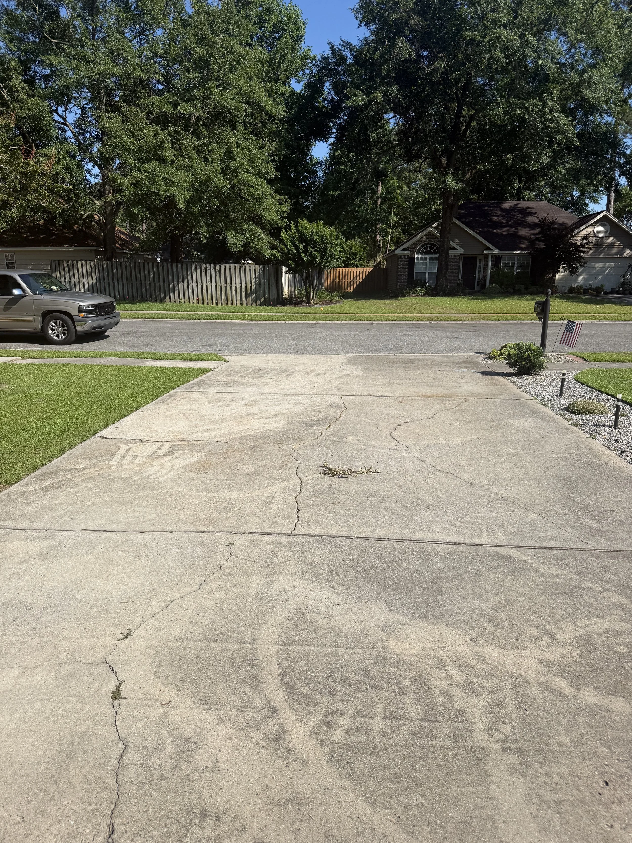 A suburban street view with a residential house, trees, and a parked car. The driveway is cracked and there is a mailbox with an American flag near a small bush.