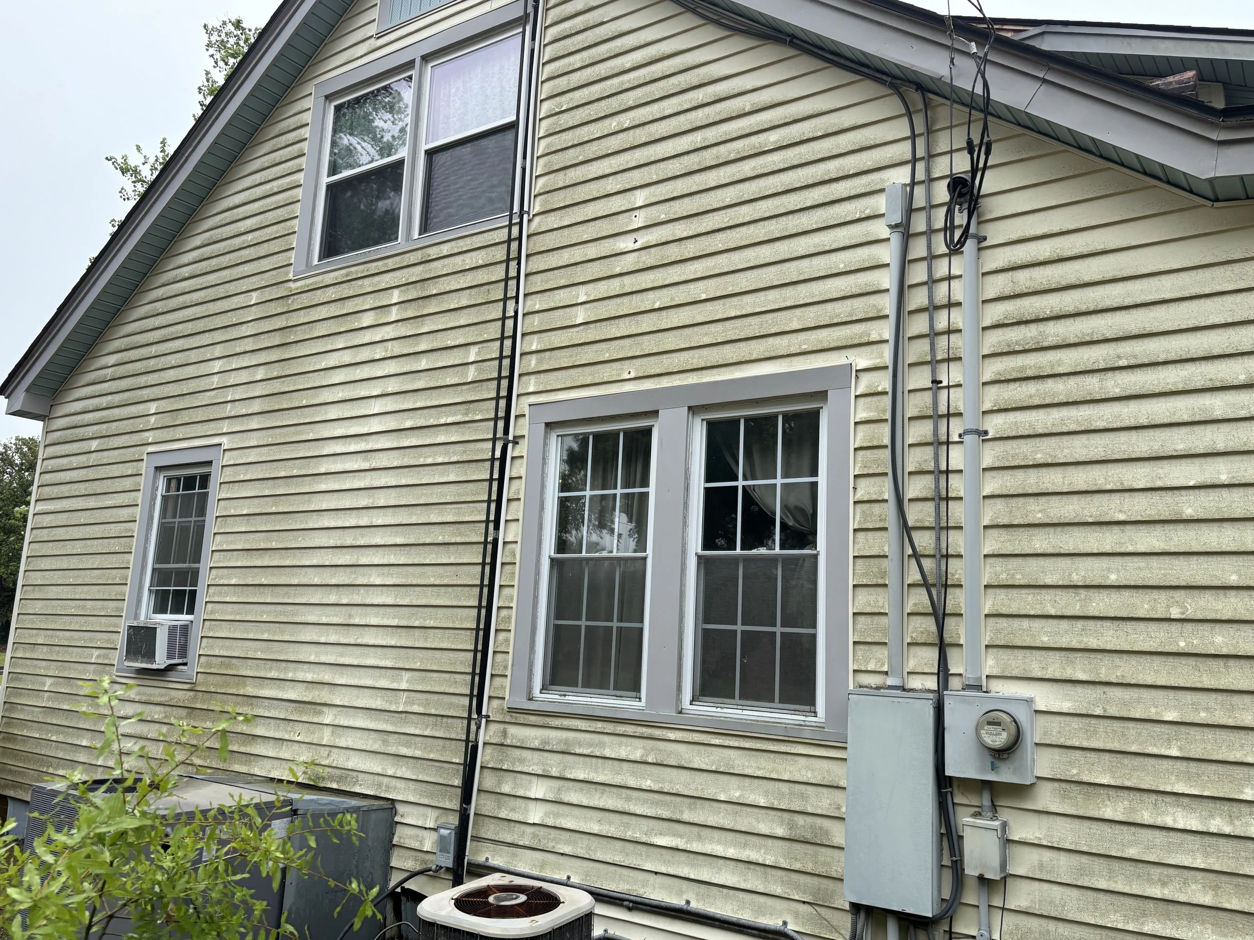 The image shows the exterior of a house with yellow siding, two white-framed windows, and various black and gray utility wires running along the wall. An air conditioning unit is installed below one of the windows, and an electric meter is visible on