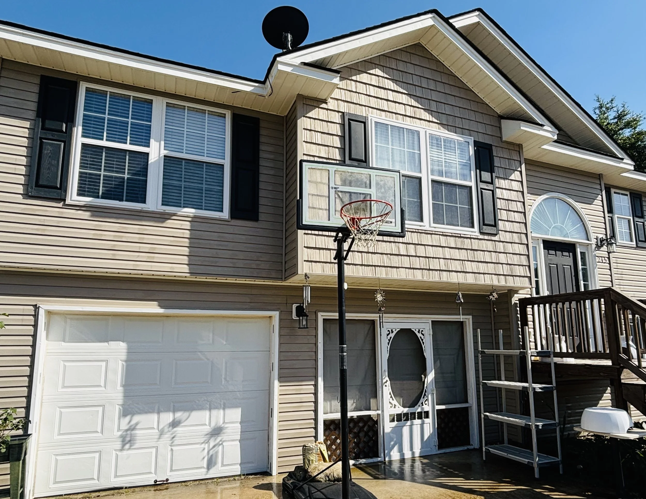 Front view of a two-story house with beige siding, multiple windows, a garage door, a basketball hoop, and outdoor decorations under a clear blue sky.