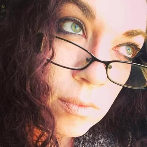 Close-up of a woman with curly purple hair and green eyes wearing black-rimmed glasses, looking to the side with a neutral expression.