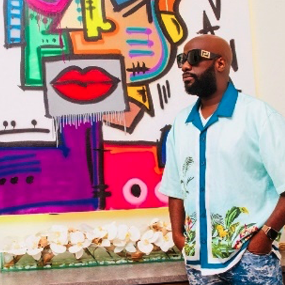 A man with a beard and sunglasses standing in front of a colorful graffiti mural featuring abstract shapes and red lips, next to a row of white orchids.