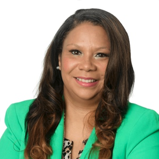 Portrait of a woman with long wavy brown hair, wearing a teal blazer and a patterned top, smiling against a white background.