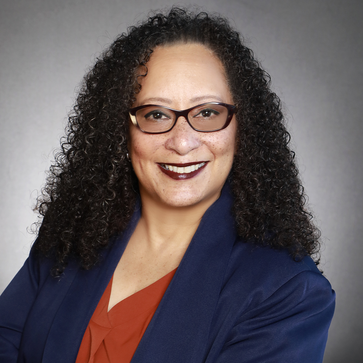 Professional portrait woman with curly dark hair, wearing glasses, a navy blazer, and a rust-colored top, smiling against a gray background.