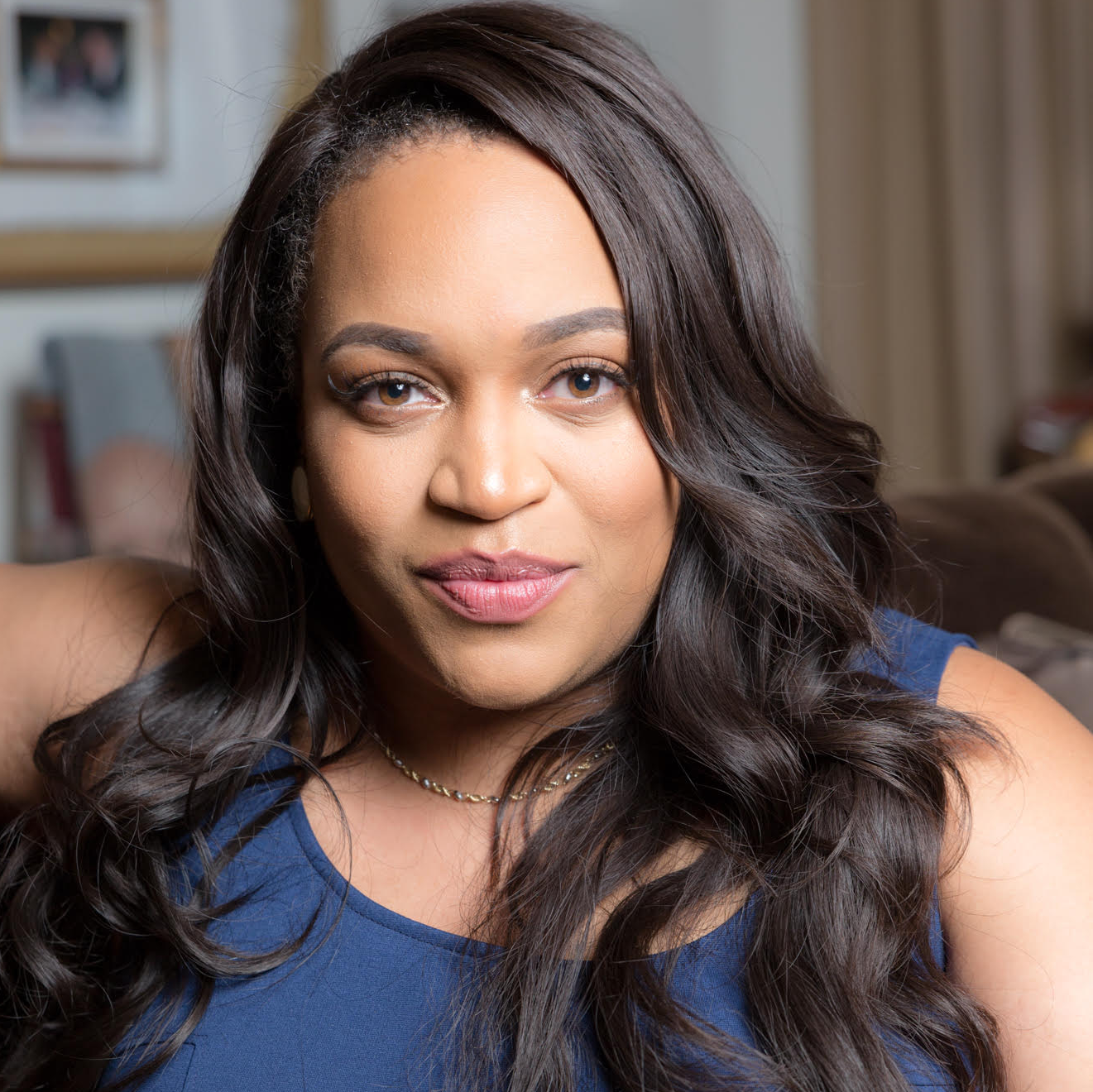 Close-up portrait of a woman with long dark wavy hair wearing a blue top, sitting in a cozy indoor setting.