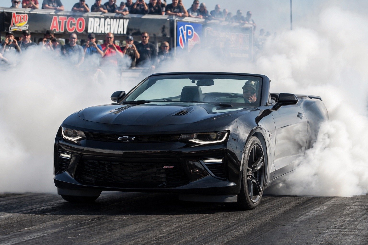 A black Chevrolet Camaro convertible performing a burnout on a drag strip, producing smoke with a crowd of spectators watching in the background at Atco Dragway.