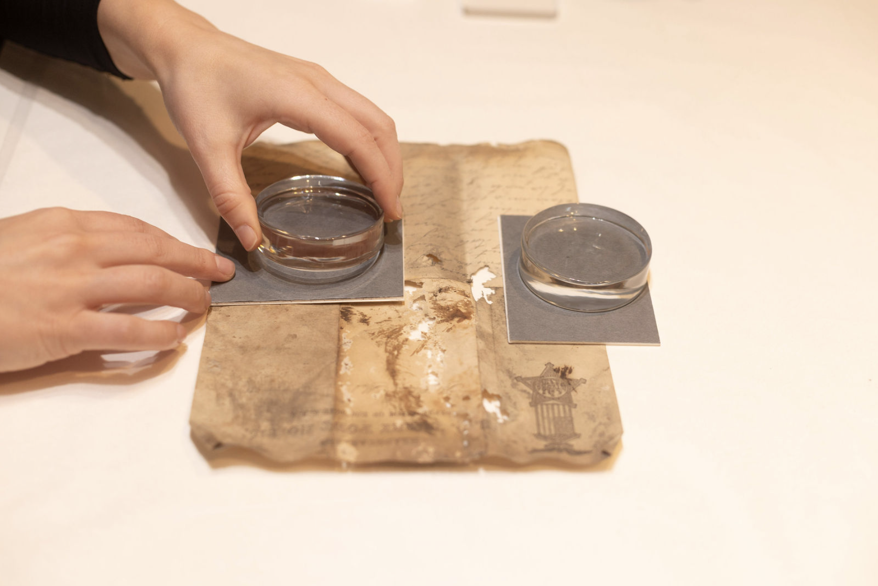 A person's hands carefully placing a glass weight on an aged, worn piece of paper with handwritten notes.