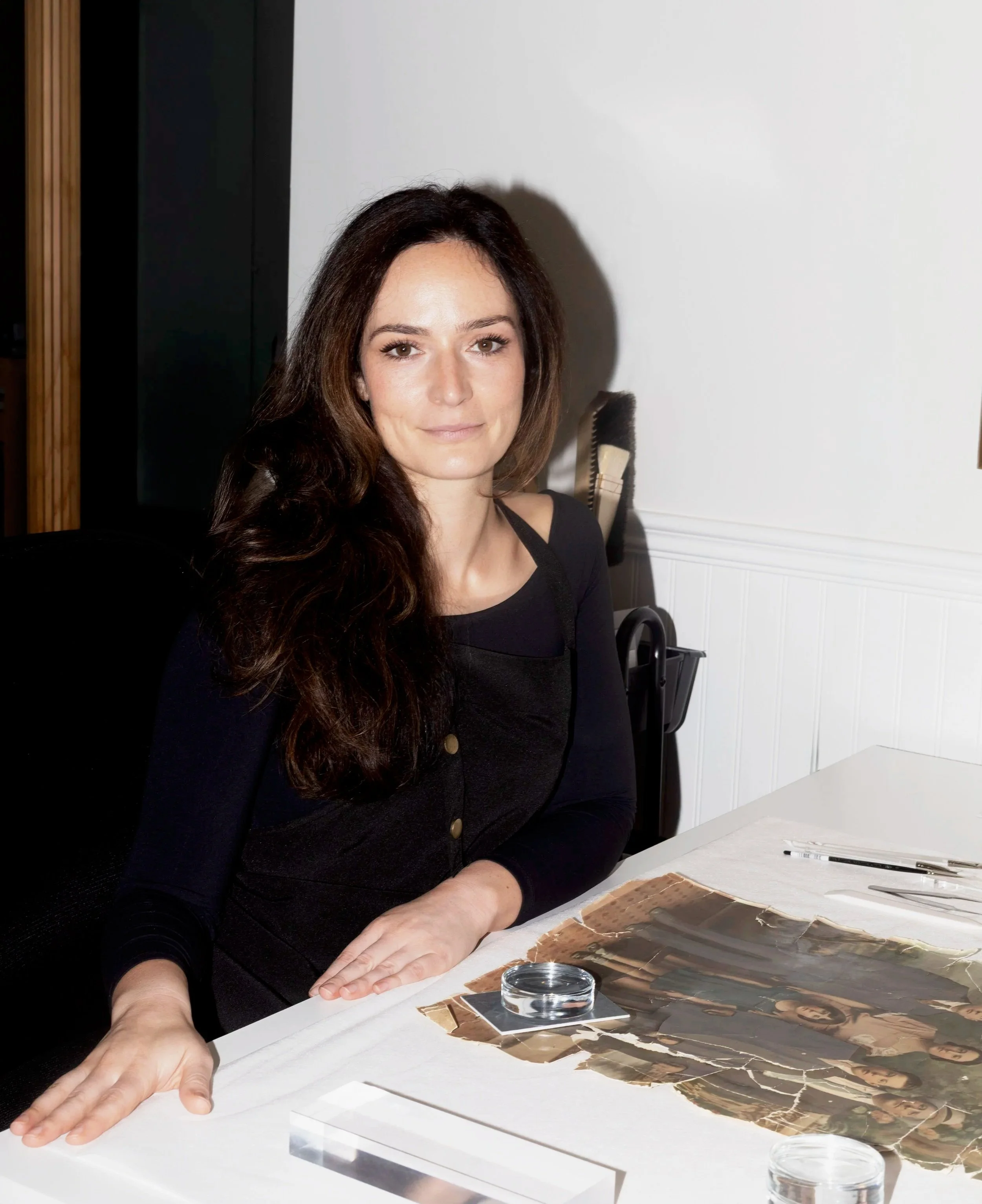 A woman with long brown hair and a black shirt sits at a white table examining a large photograph with brushes and tools nearby.
