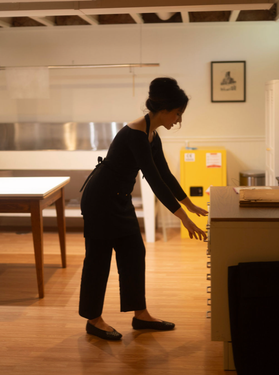 A woman dressed in black, bending forward to open or close a drawer in a conservation studio.
