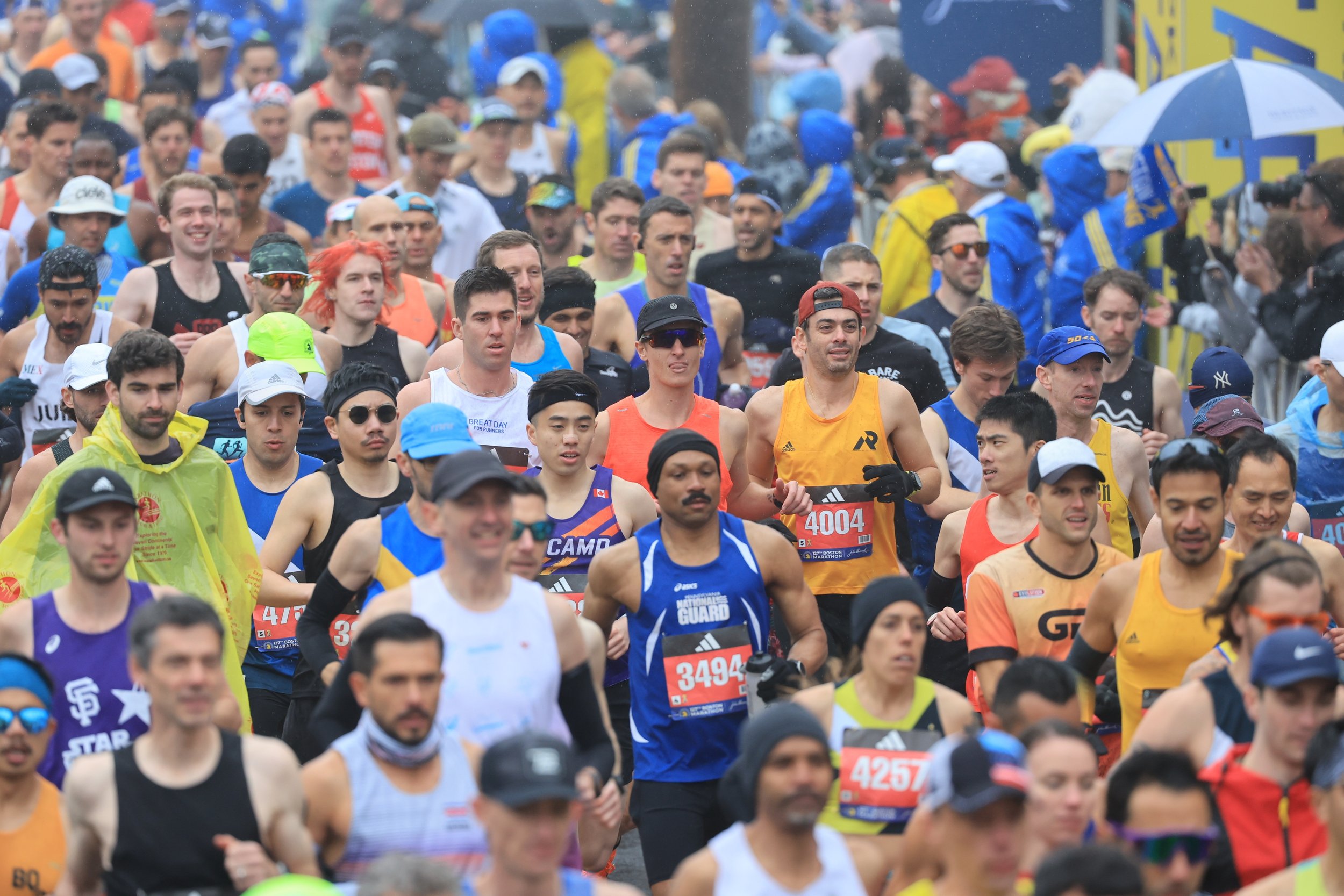 A large group of runners participating in a marathon race outdoors, with some wearing colorful athletic gear and bib numbers, rain visible, and various spectators or race officials in the background.