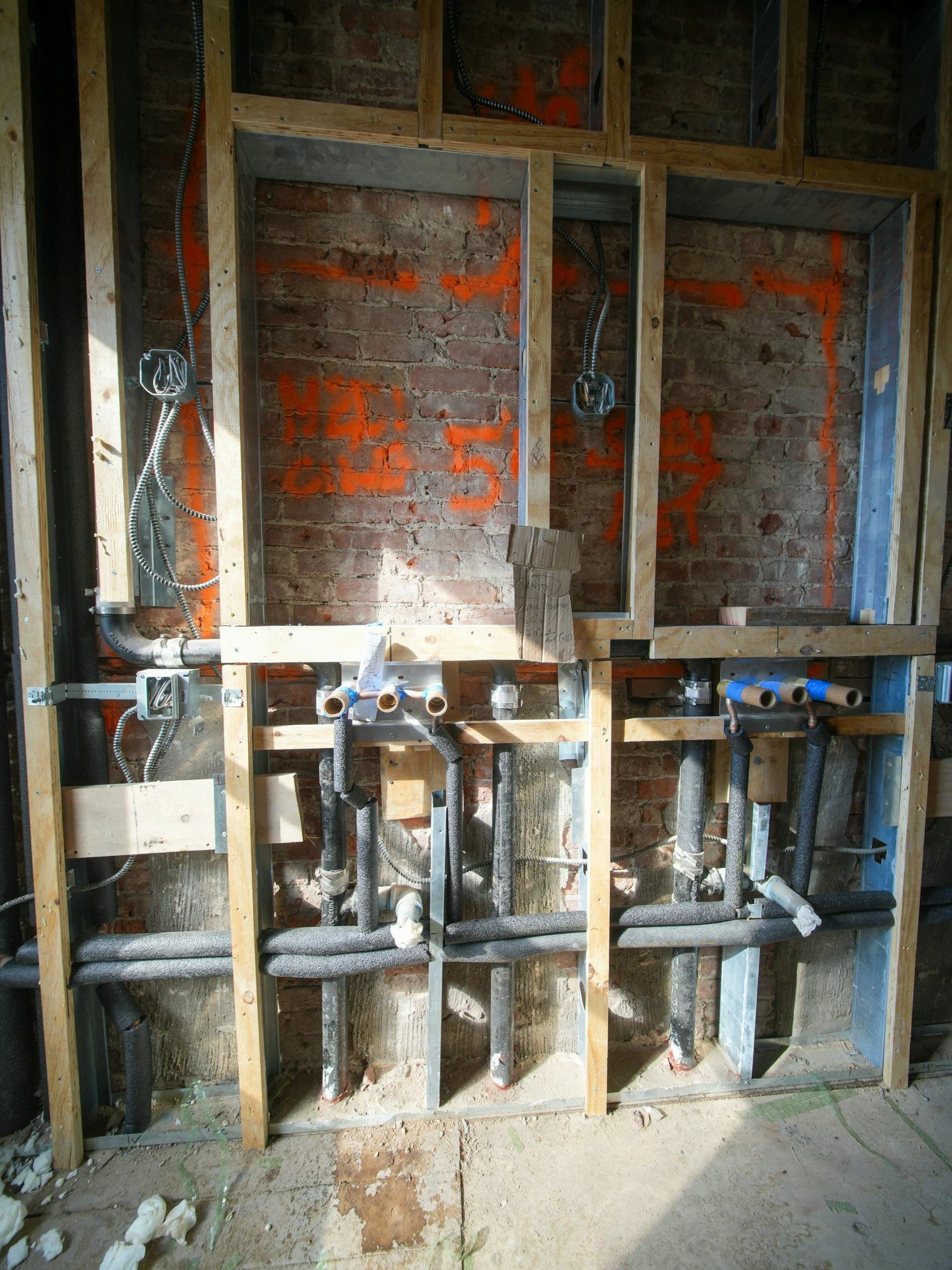 Construction site with exposed brick wall and plumbing pipes, electrical wiring, and wooden framework for walls.