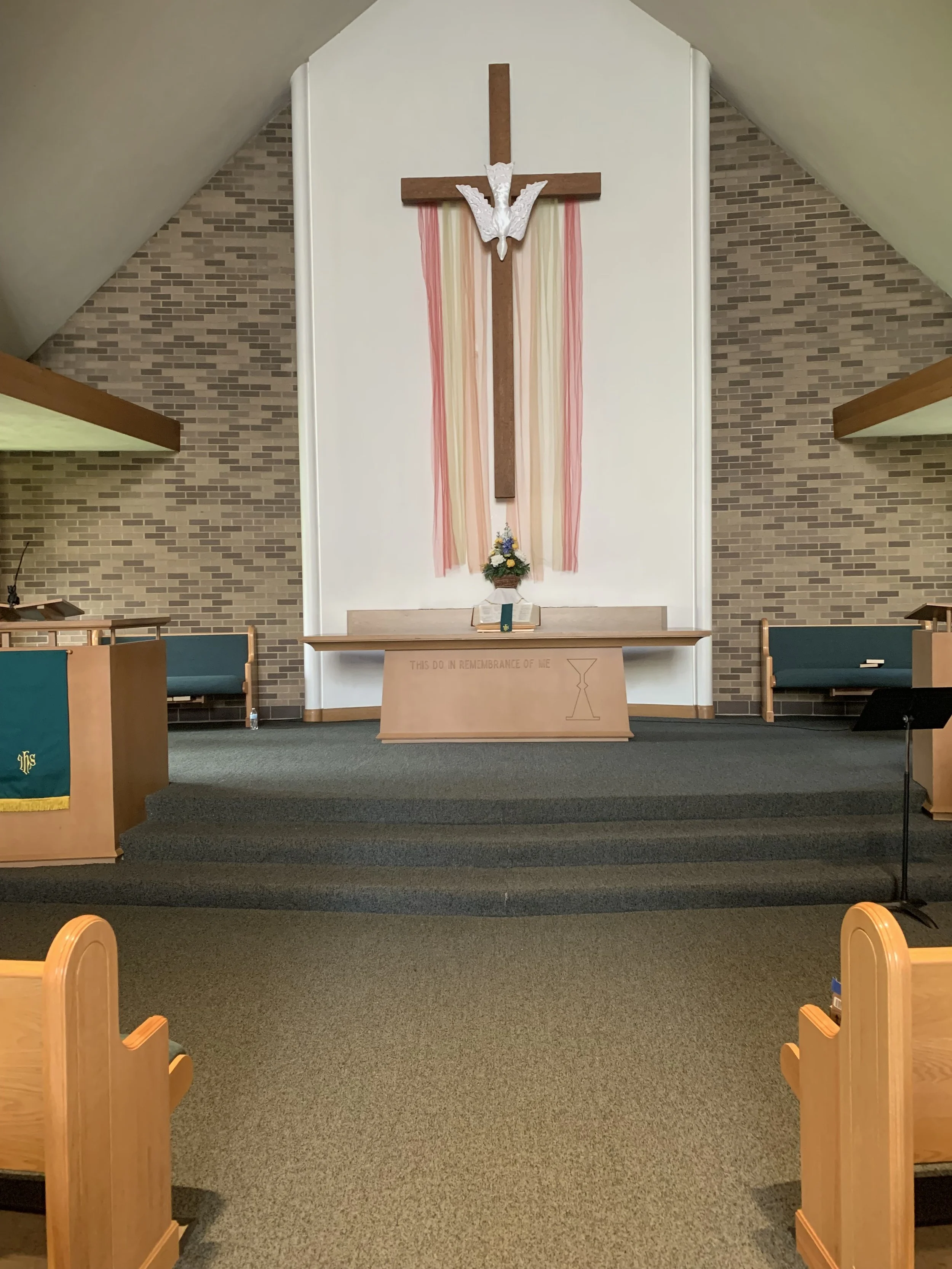 Inside a church, view of the altar with a wooden cross, adorned with a white dove and pink, beige, and white streamers. There are pews and hymnals around.