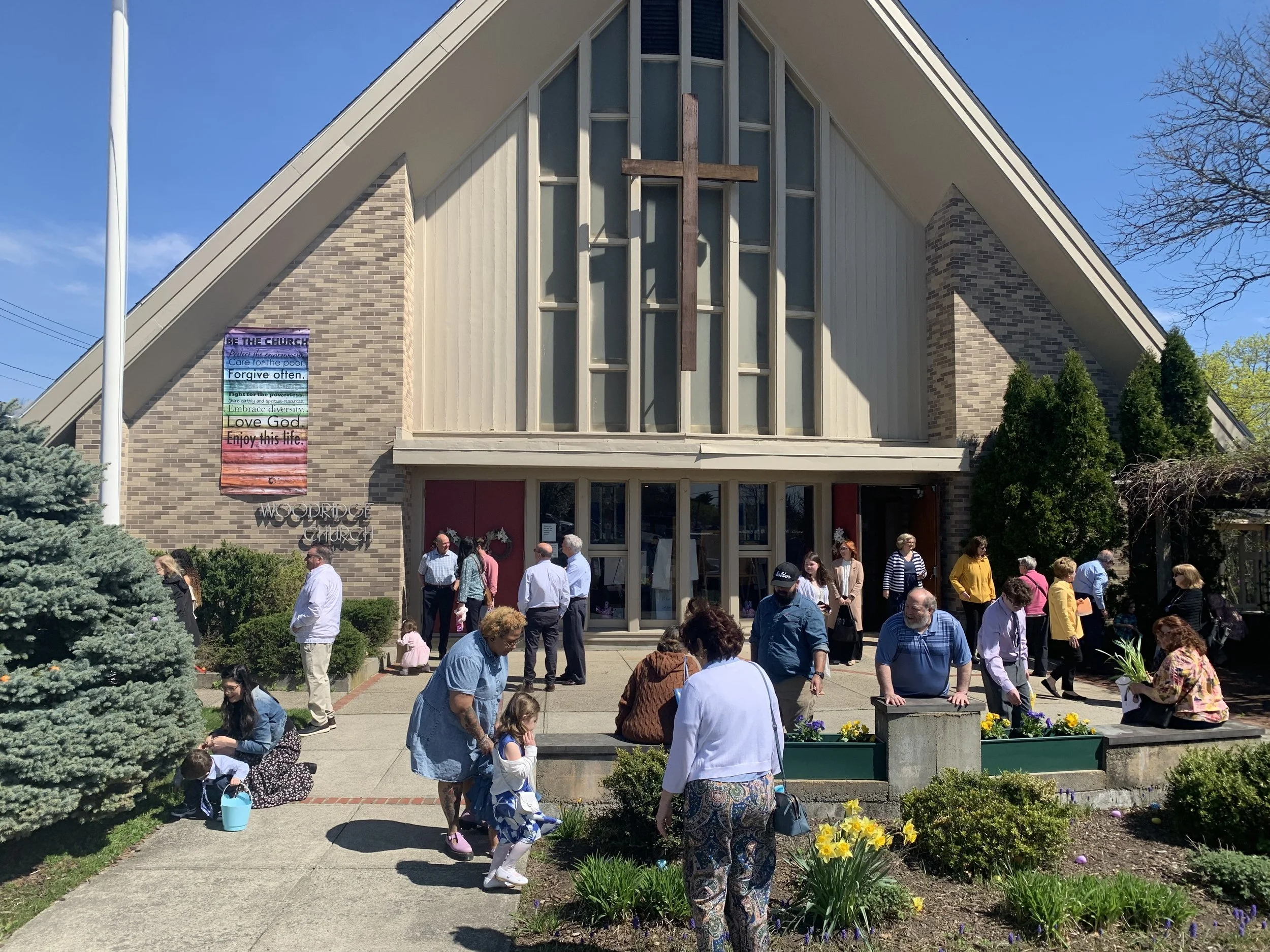 People gathered outside Woodridge Church on a sunny day, socializing and children playing in a garden with blooming flowers in front of a modern church building with a prominent cross and windows.