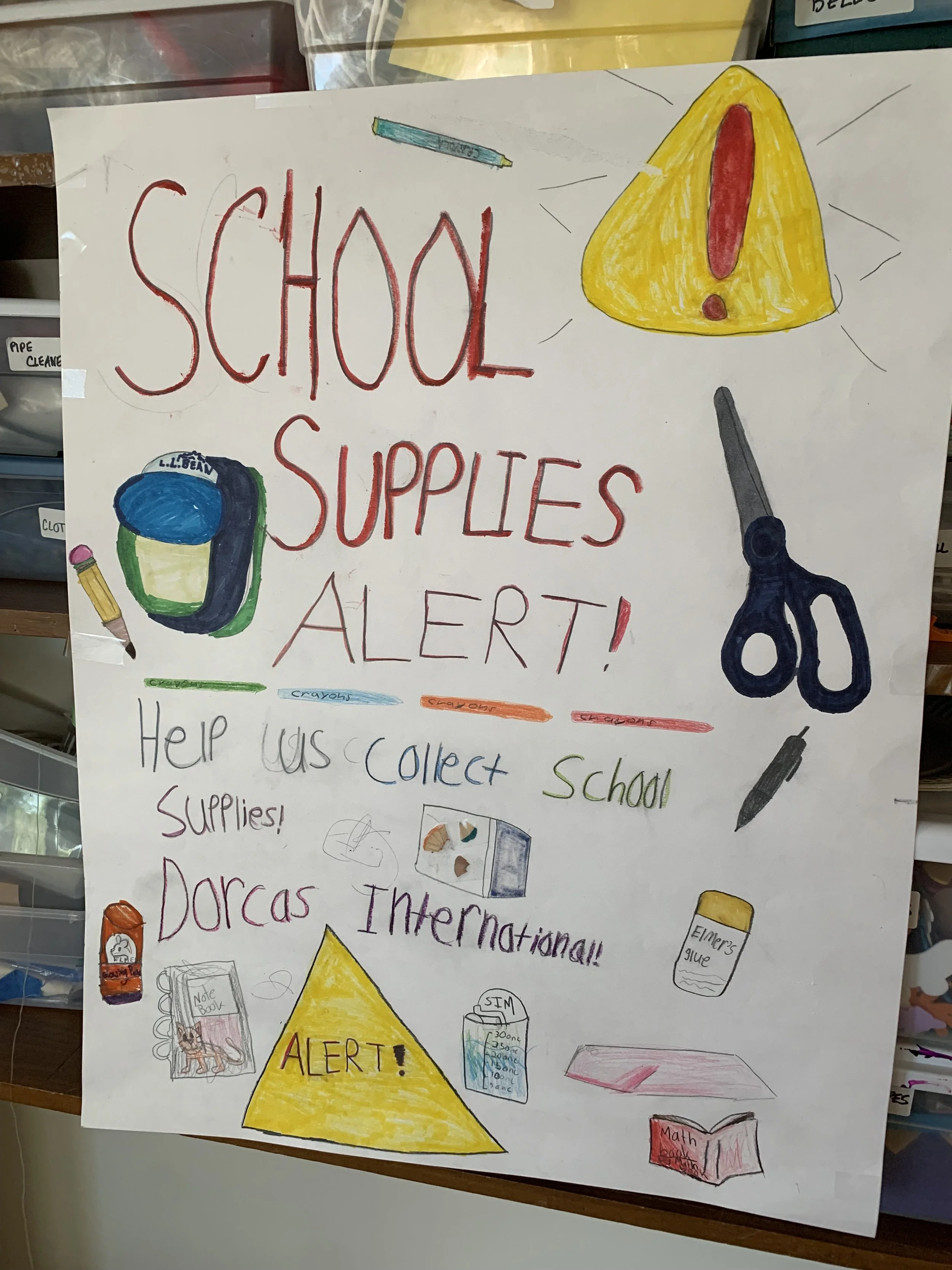 A hand-drawn sign on white paper with the words 'SHOOL SUPPLIES ALERT!' in red and black, accompanied by colorful drawings of school supplies including a pencil, a backpack, a pair of scissors, a glue bottle, a notebook, and a marker, alerting about school supplies.