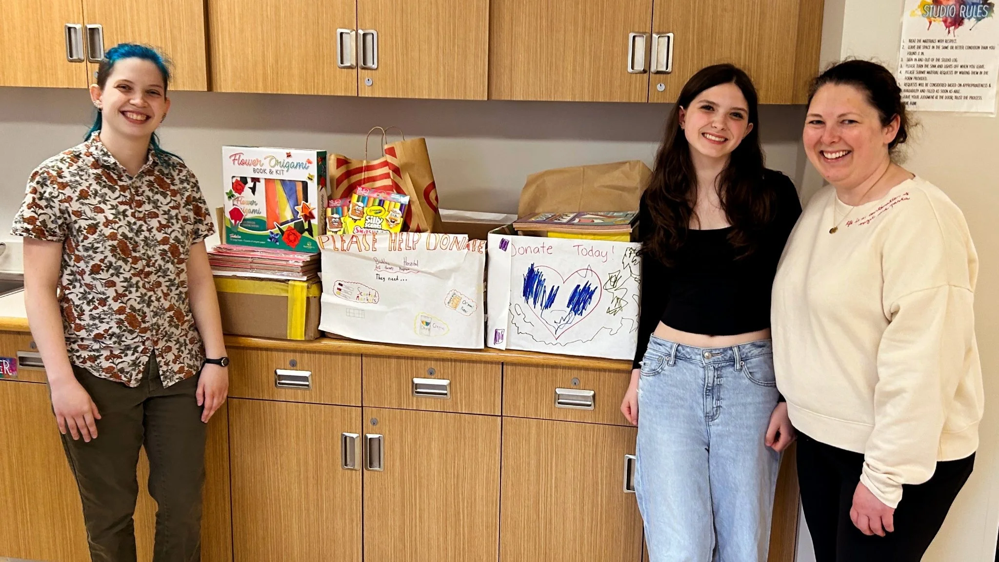 Three women standing in front of a table with donation boxes and gifts, smiling at the camera.