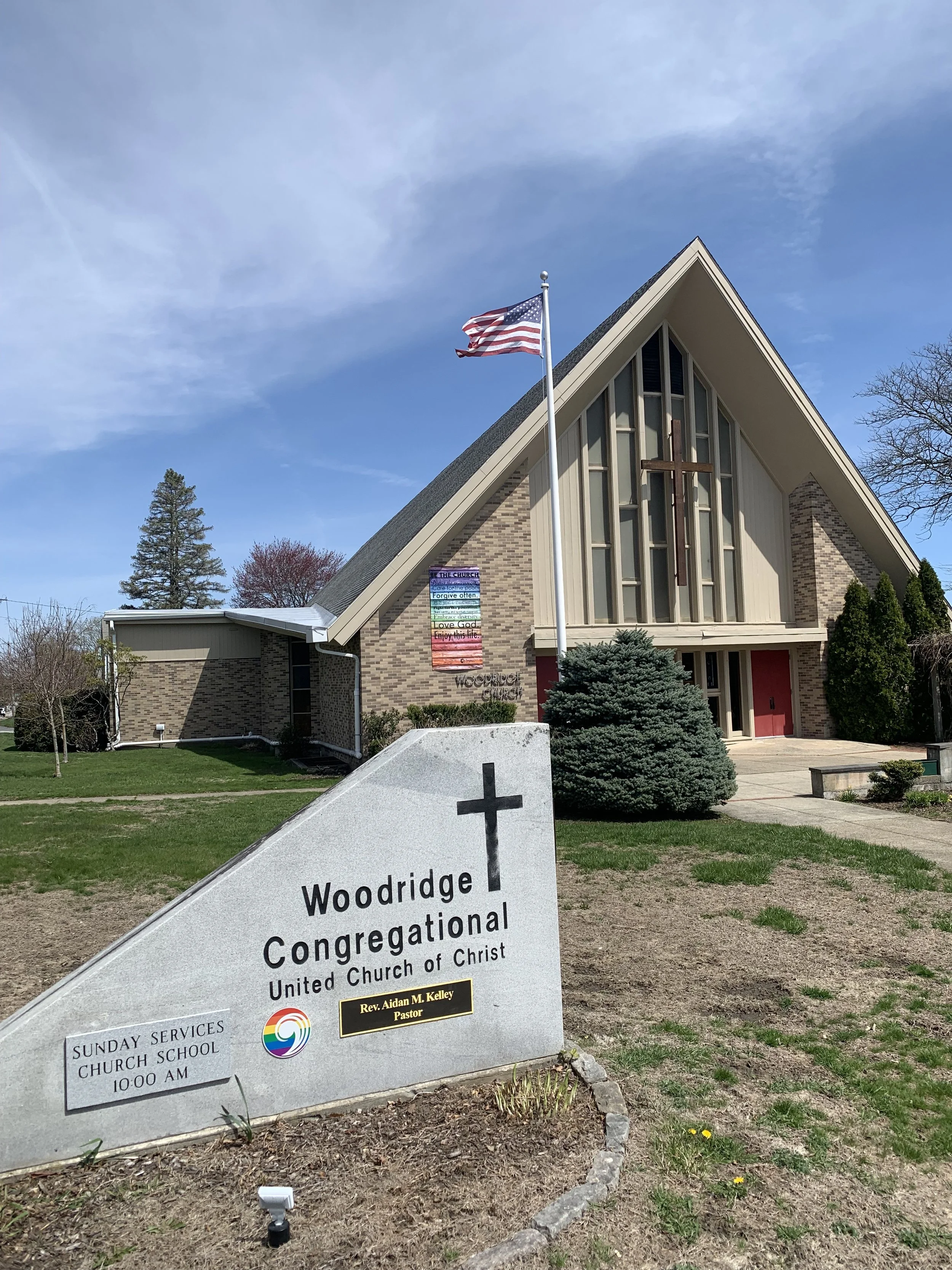 Sign in front of a church with the text "Woodridge Congregational United Church of Christ" and a cross logo. The church building has modern architecture with large glass windows and a steep roof, and an American flag on a flagpole. There are trees and a partly cloudy sky in the background.