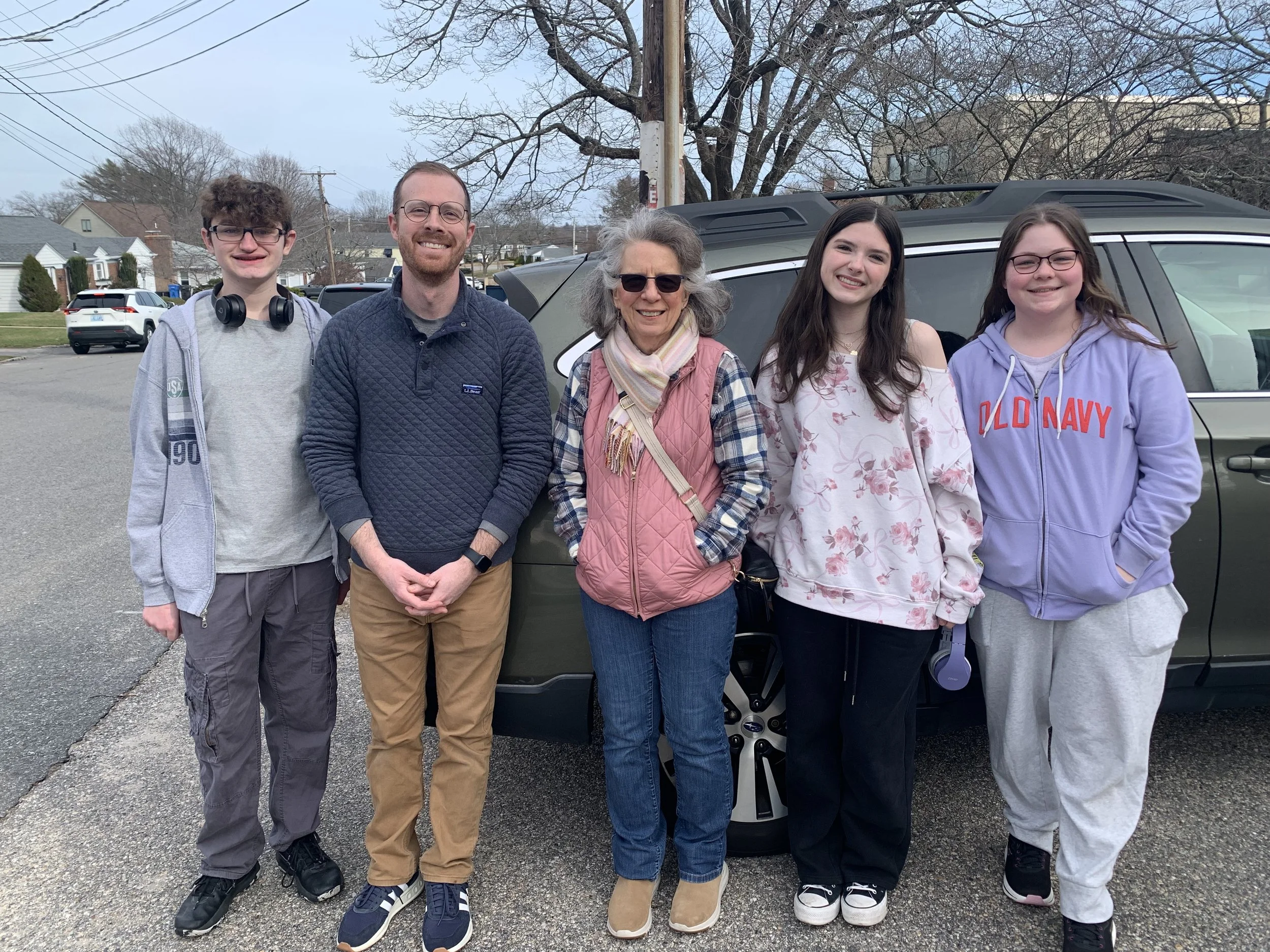 Group of five people standing outdoors in front of a car, smiling, with trees and houses in the background.