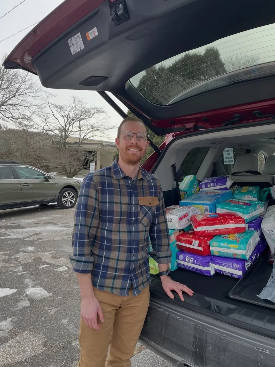 A man standing next to an open car trunk filled with packs of diapers, smiling at the camera, in a parking lot with some snow on the ground and a grey car in the background.