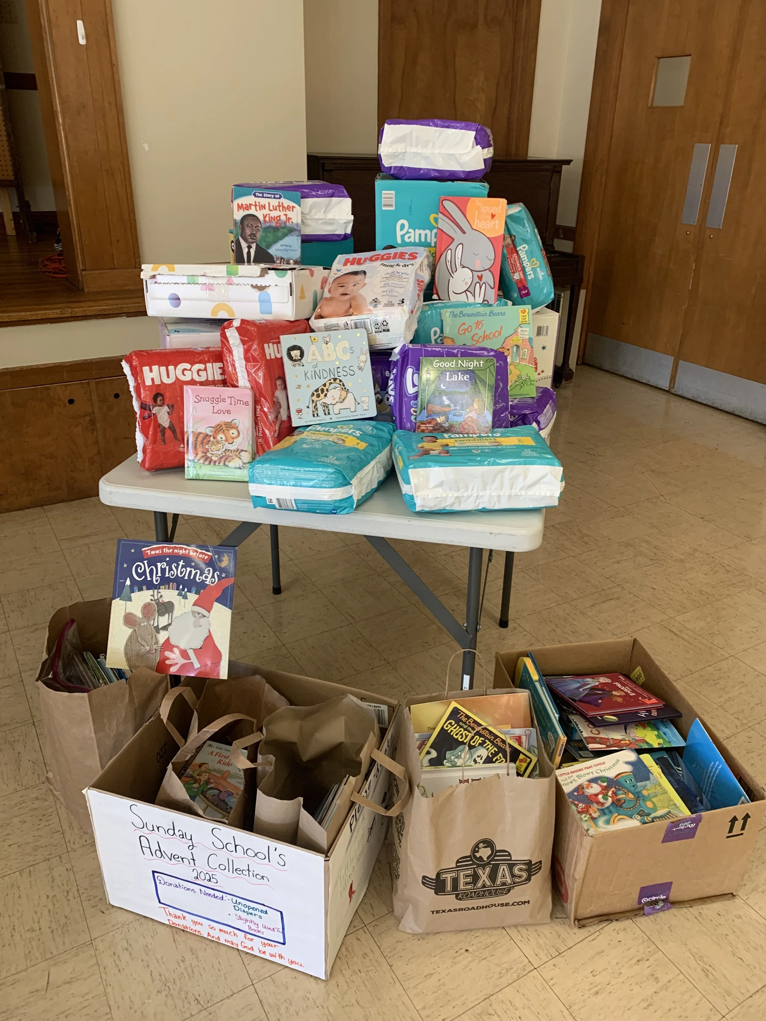 Tables filled with donated items including books, toys, and packages of diapers, with a sign that reads 'Sunday School's Advent Collection 2025' and a box of books nearby.