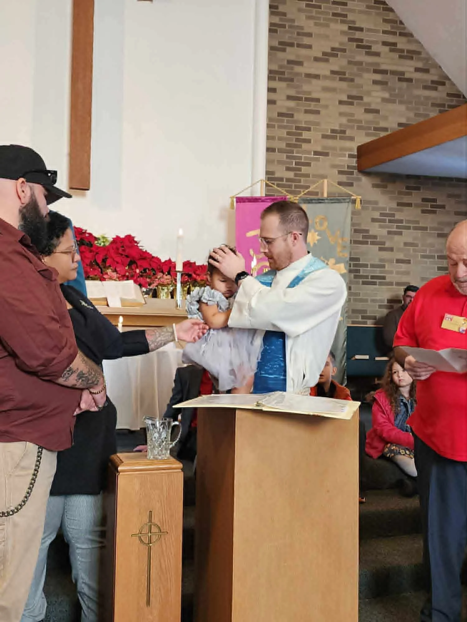 A person holding a baby during a baptism, pouring water over the baby's head from a scallop shell, with a white baptismal font in the foreground.