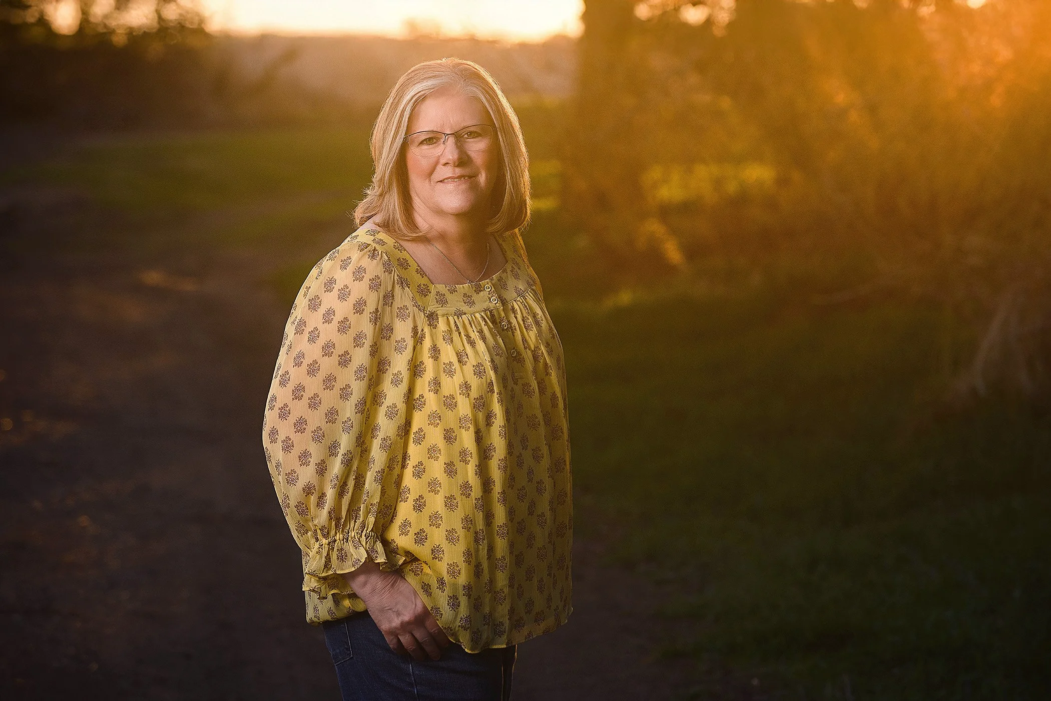 A woman with blonde hair, glasses, and a yellow patterned blouse standing outdoors during sunset, with trees and sunlight in the background.