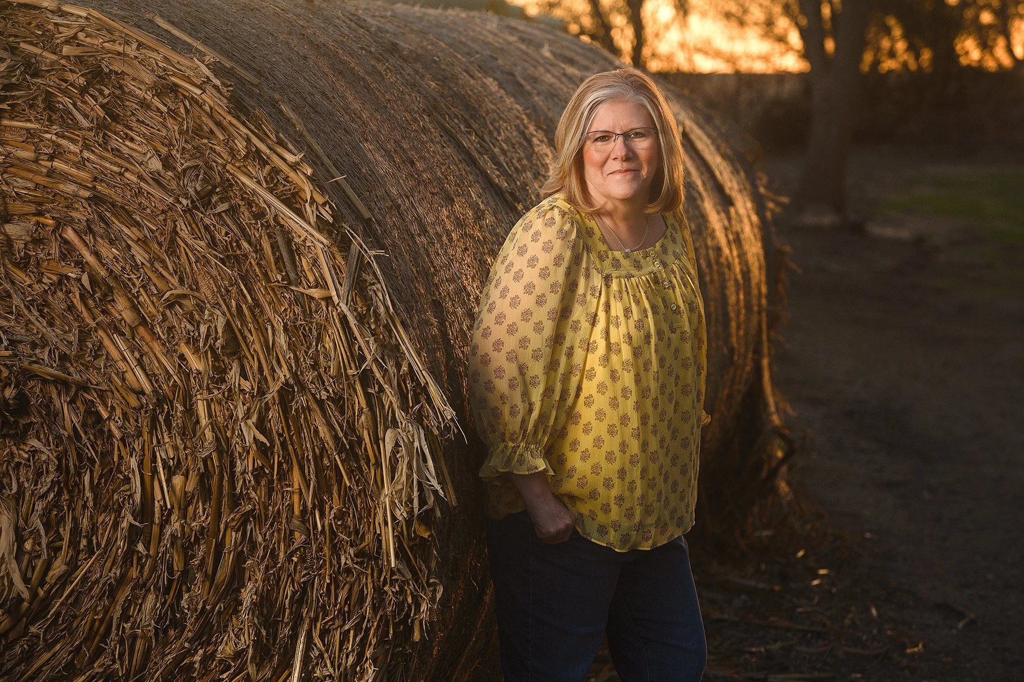A woman with glasses and shoulder-length blonde hair stands beside a large hay bale during sunset. She is wearing a yellow dress with a floral pattern and black pants, looking directly at the camera with a slight smile.
