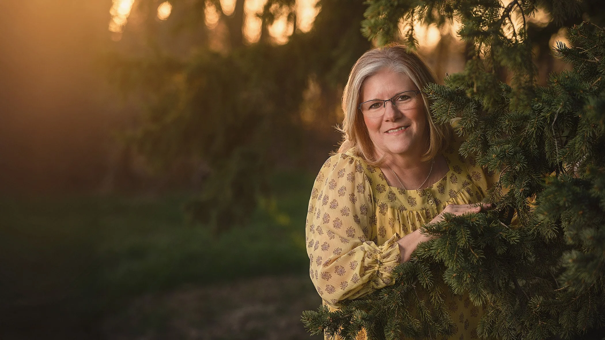 A woman with gray hair and glasses smiling as she peeks from behind a pine tree during sunset.