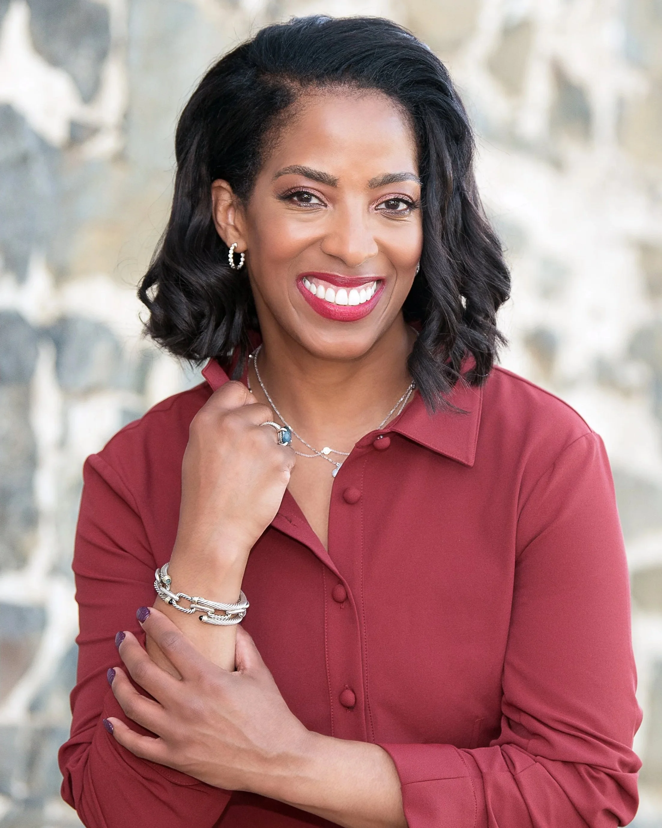 A woman with dark, wavy shoulder-length hair smiling, wearing a red blouse and jewelry against a blurred outdoor background.