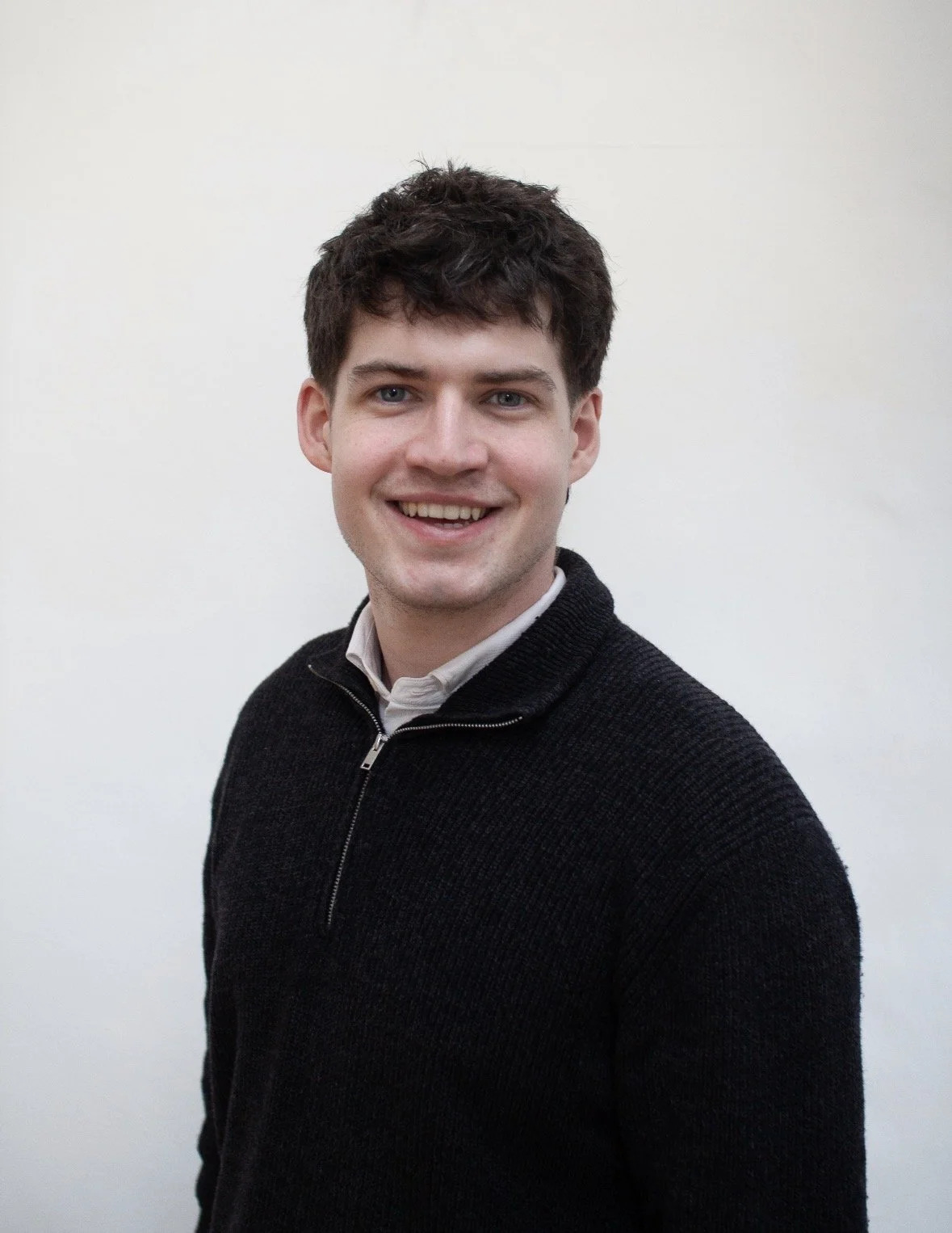 A young man with dark brown hair, wearing a black zip-up sweater over a white collared shirt, smiling against a plain white background.