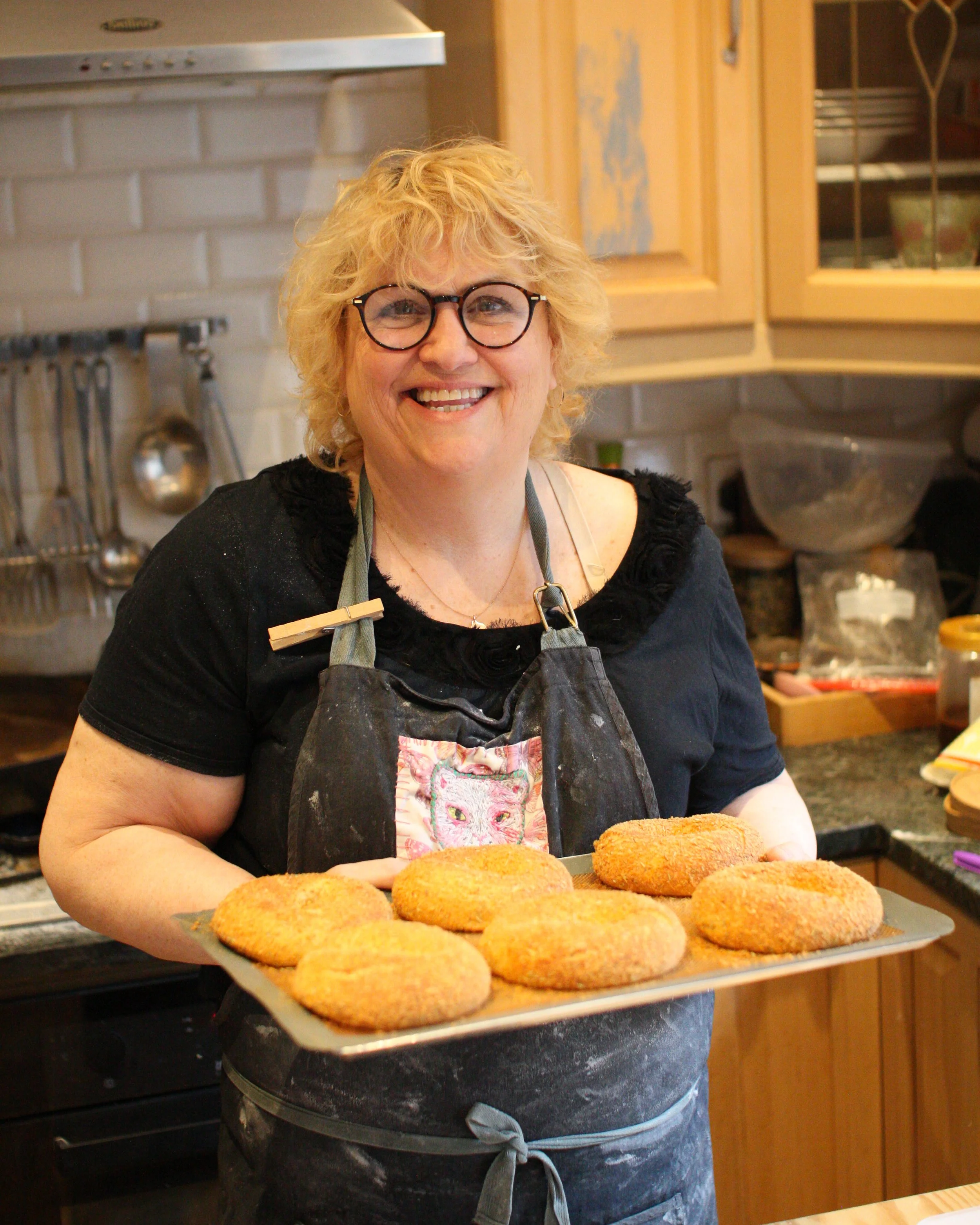 A woman with blonde curly hair and glasses smiling while holding a tray of breaded hamburgers in a kitchen.